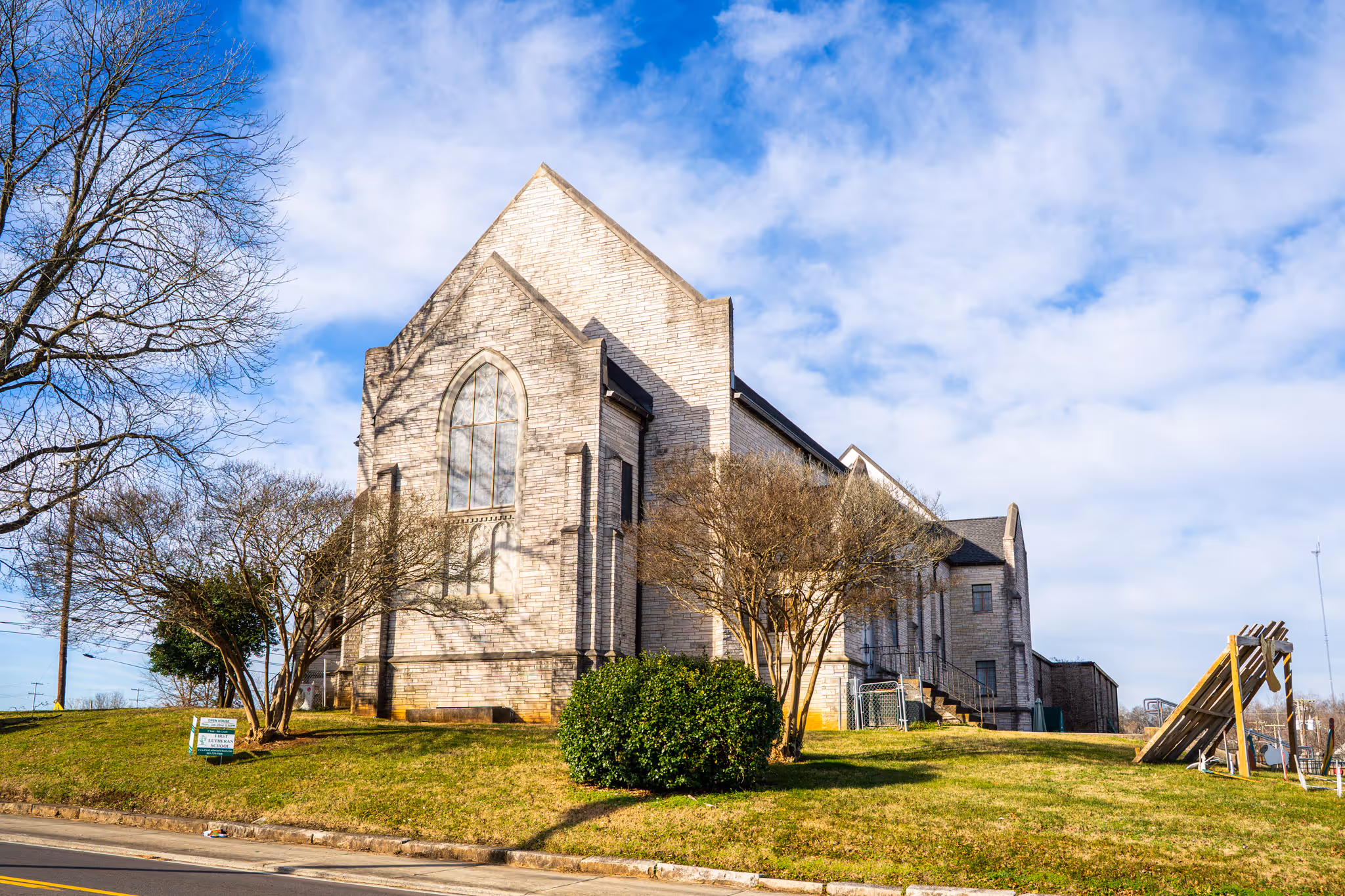 Stone church building with large arched window on a grassy hill under a blue sky with scattered clouds.