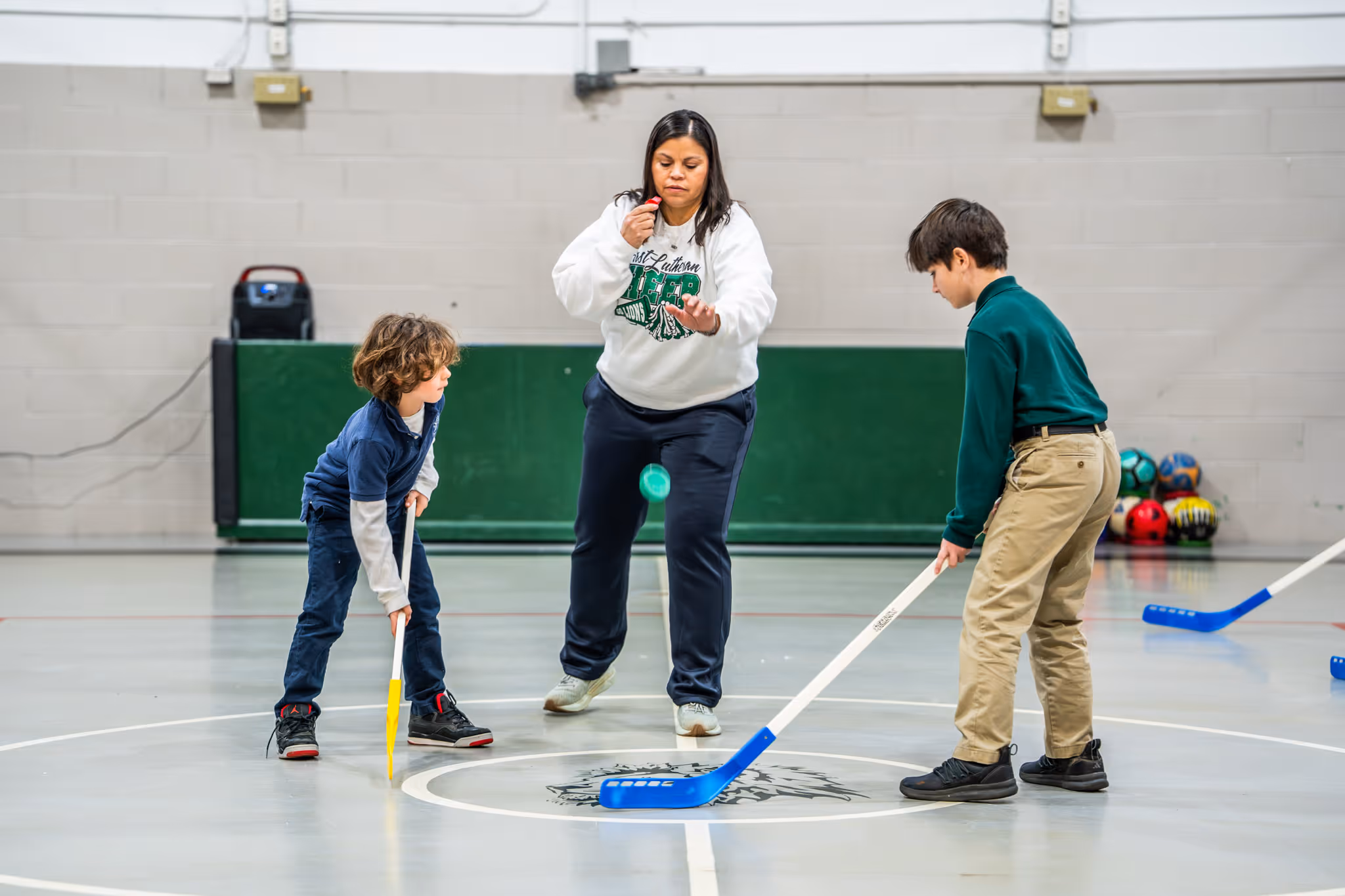 A woman wearing a whistle prepares to drop a ball between two boys holding floor hockey sticks in a gym.