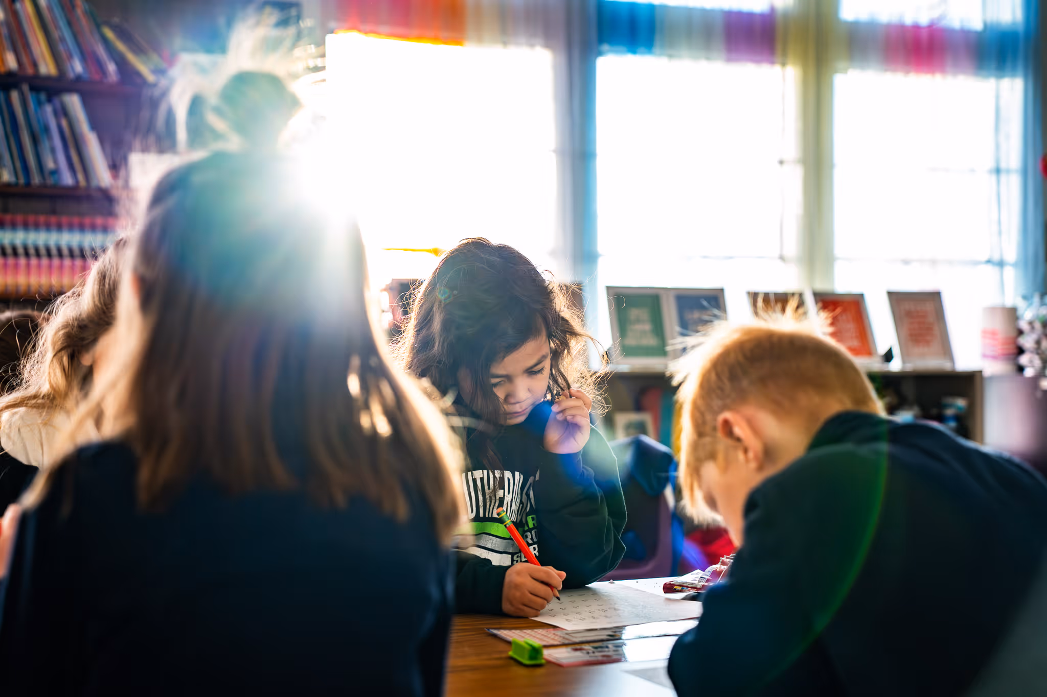 Children focused on writing at a school desk with sunlight streaming through classroom windows.