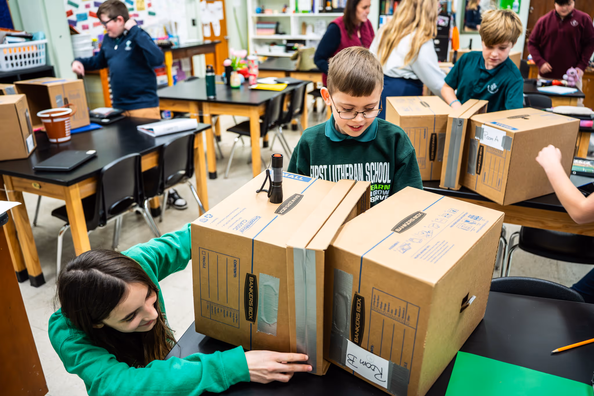 Elementary school students in a classroom building a project using labeled cardboard boxes, one labeled Room B.