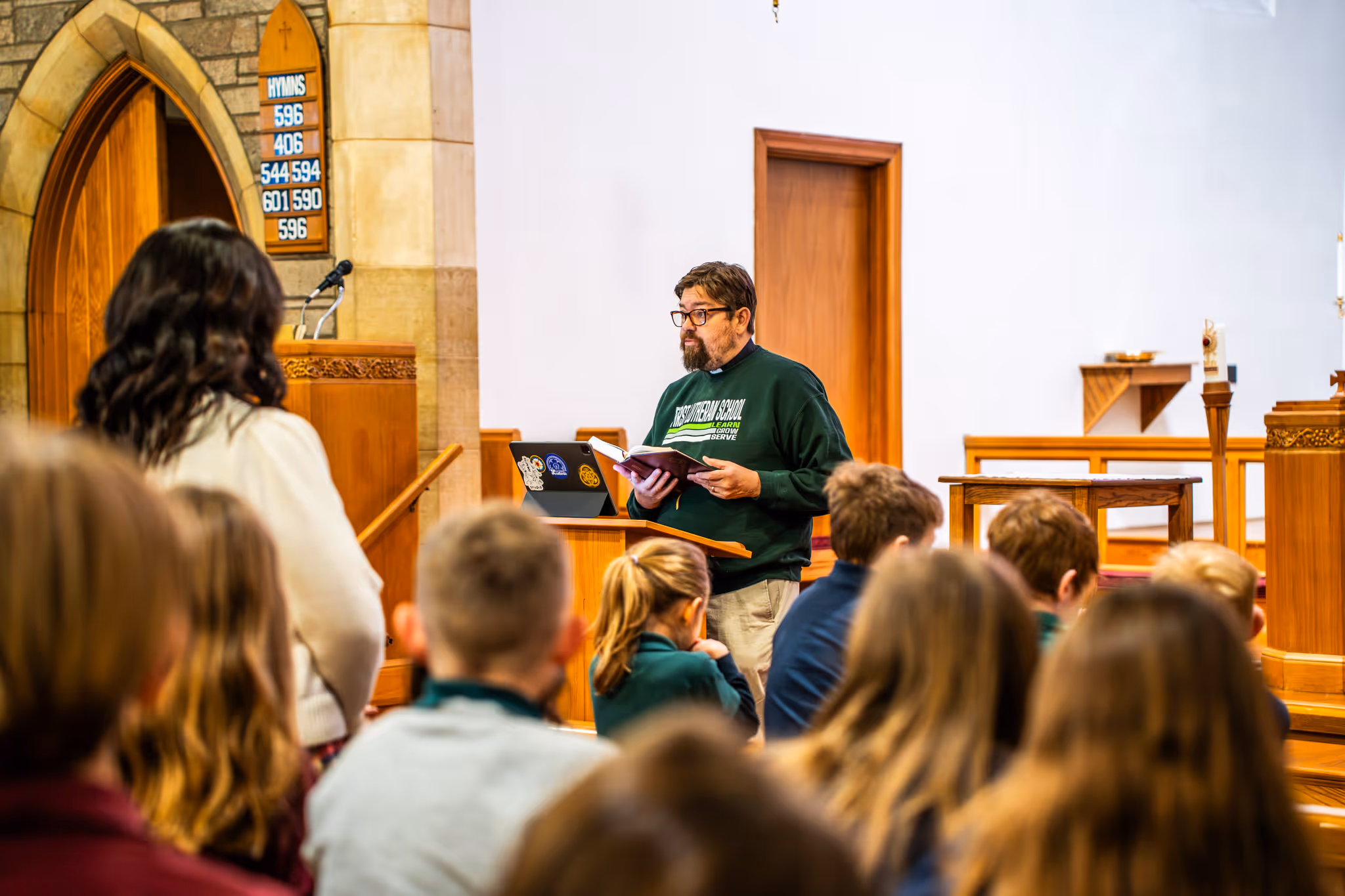 Man standing at a pulpit reading from a book in a church with children seated in front of him.