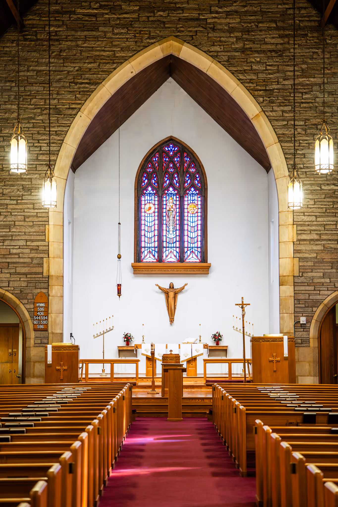 Interior of a church altar with wooden pews, a stained glass window, and a statue of Jesus with outstretched arms.