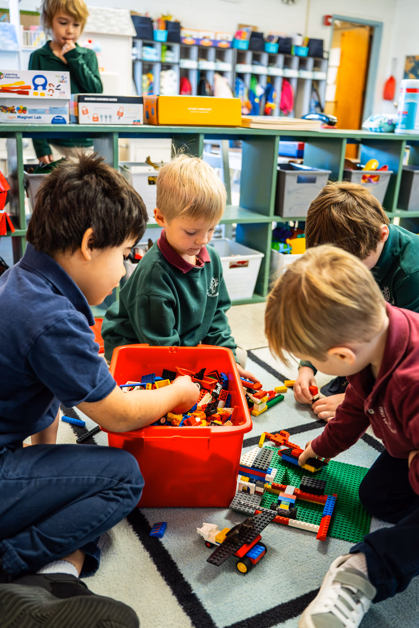Four young boys sitting on a classroom floor playing with colorful building blocks from a red container.