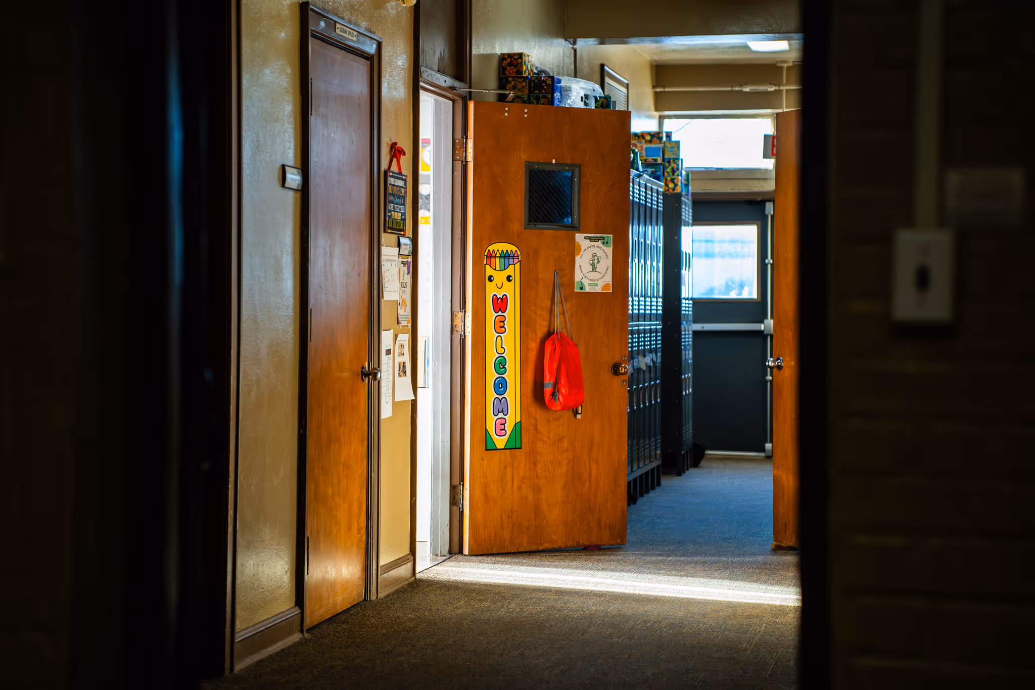 School hallway with wooden doors, one open door displaying a colorful 'Welcome' sign, lockers in the background, and a red bag hanging on the door handle.
