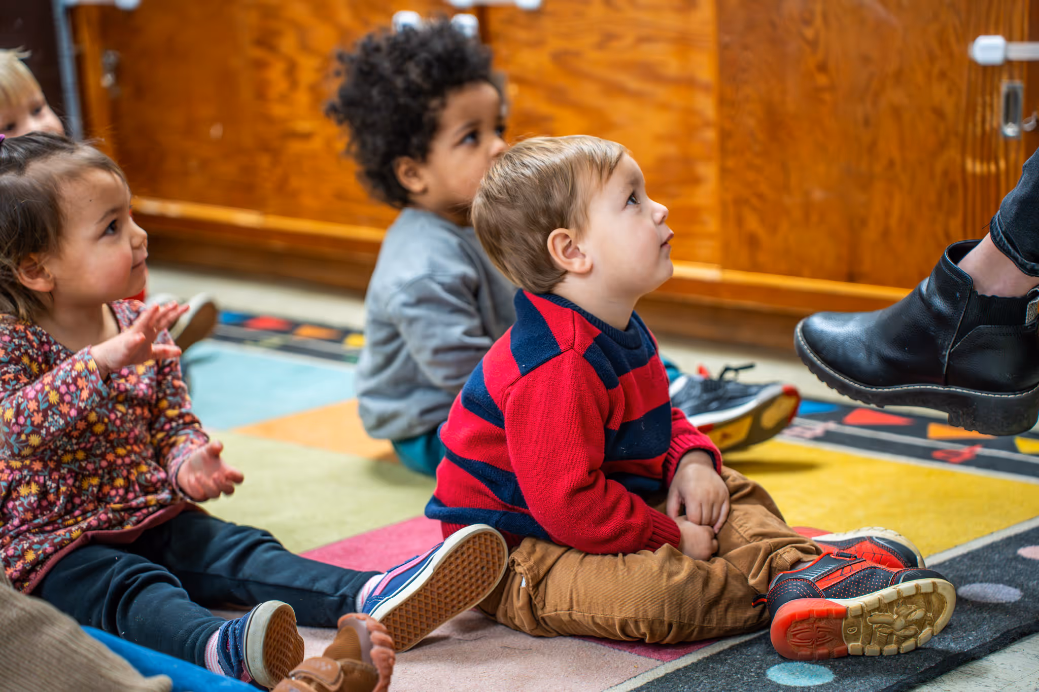 Group of young children sitting on a colorful rug looking attentively at an adult wearing black boots.