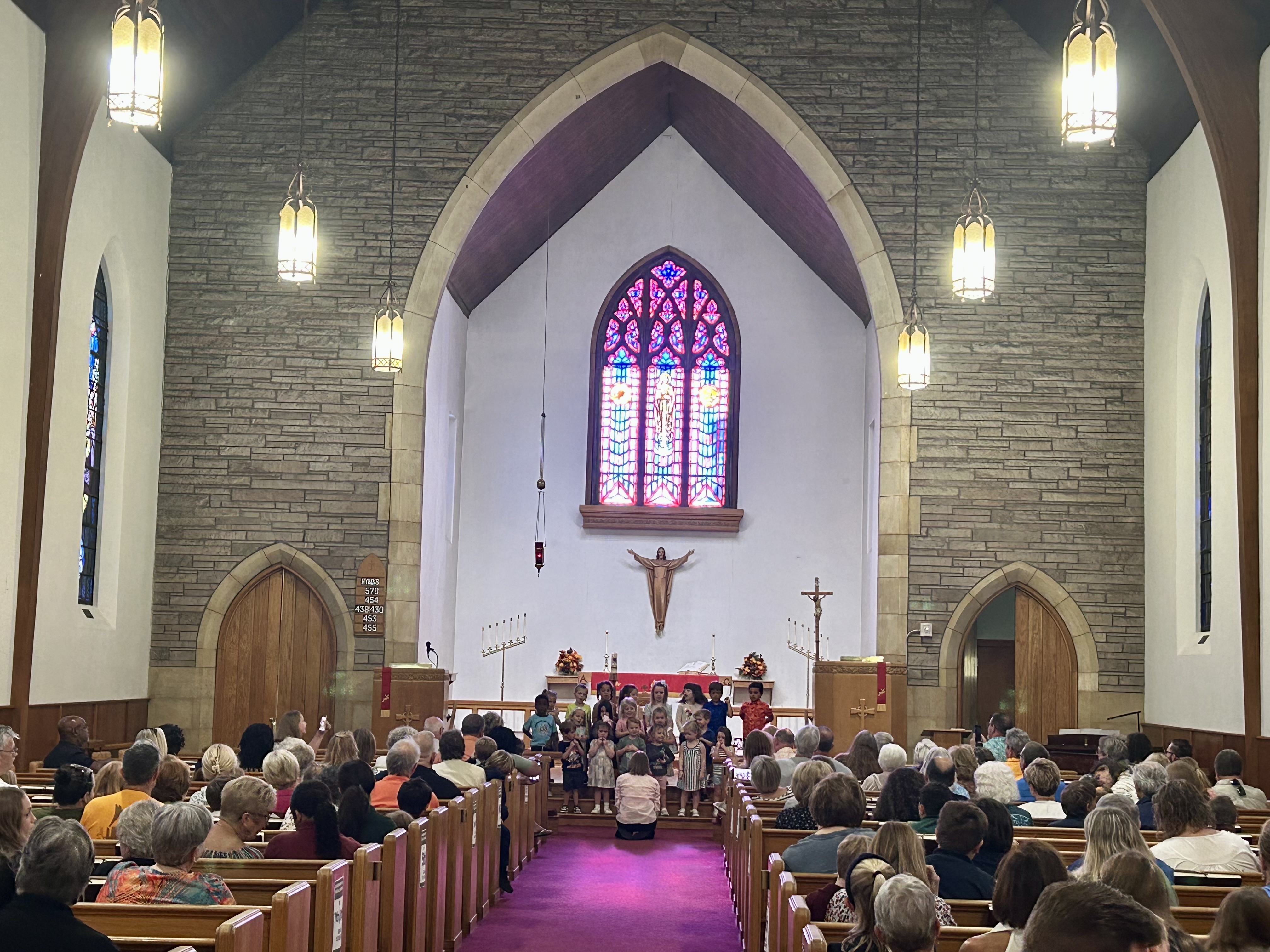 Group of smiling elementary school children sitting on wooden benches inside a classroom or assembly hall.