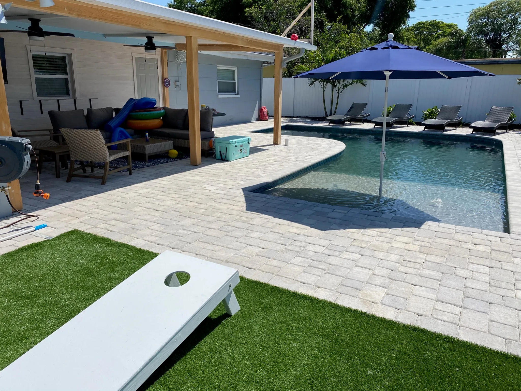 Backyard with a pool, sun loungers, a blue umbrella, patio seating area, and a cornhole board on artificial grass.