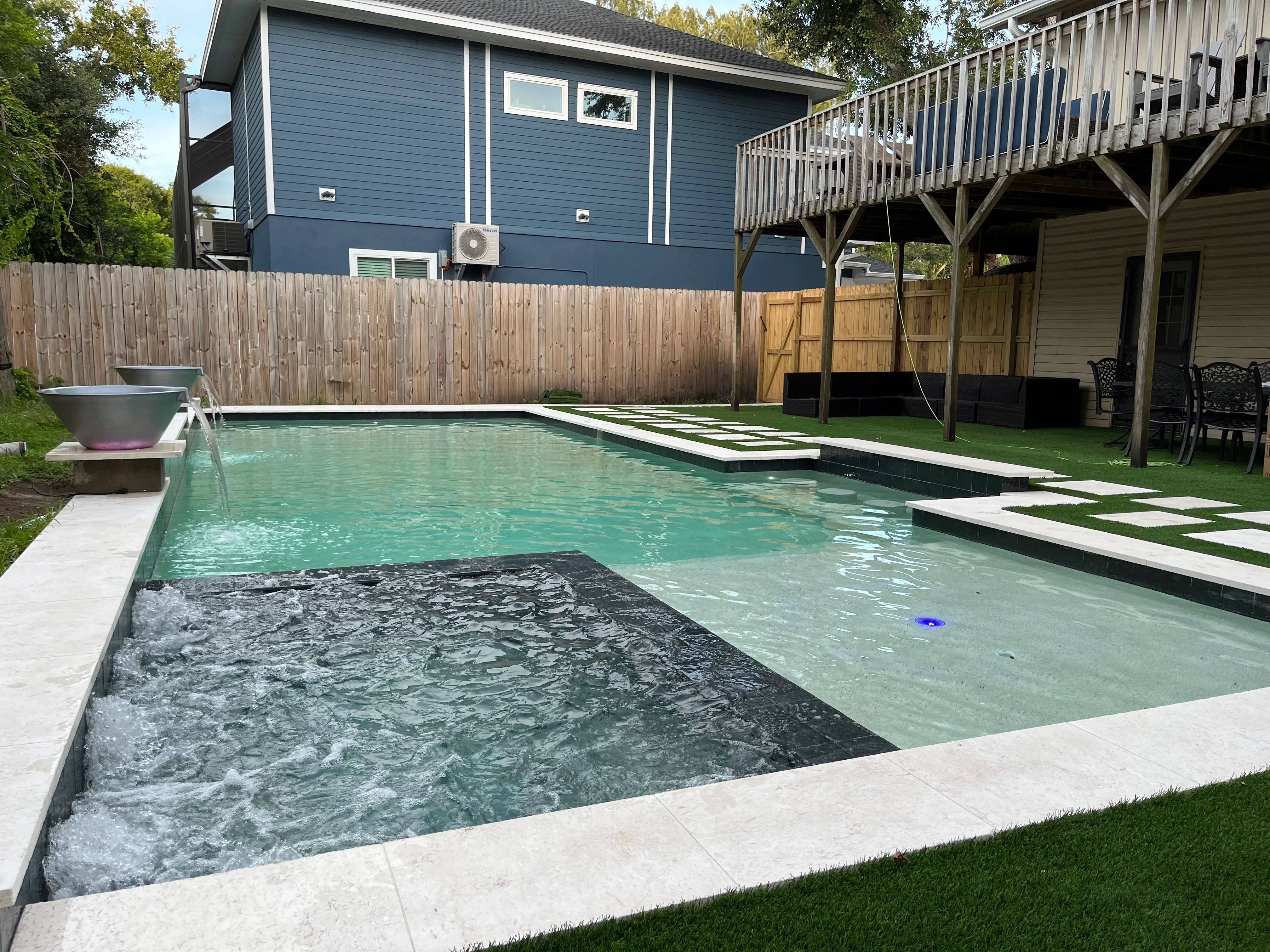 Modern backyard swimming pool with a built-in spa, textured water features, and surrounding artificial grass and patio furniture.