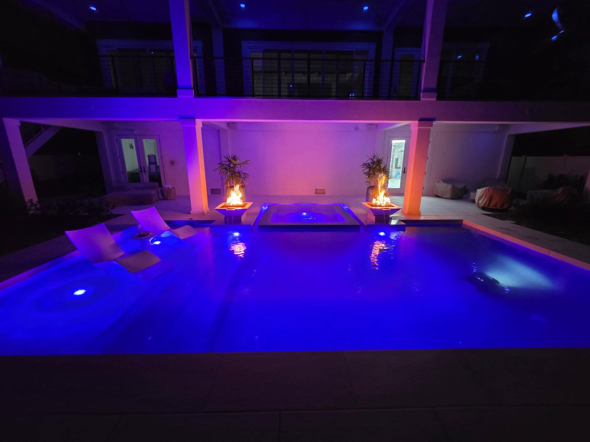 Finished YOUtopia pool at night with blue lighting, a raised spa, fire bowls and loungers on a shallow shelf.