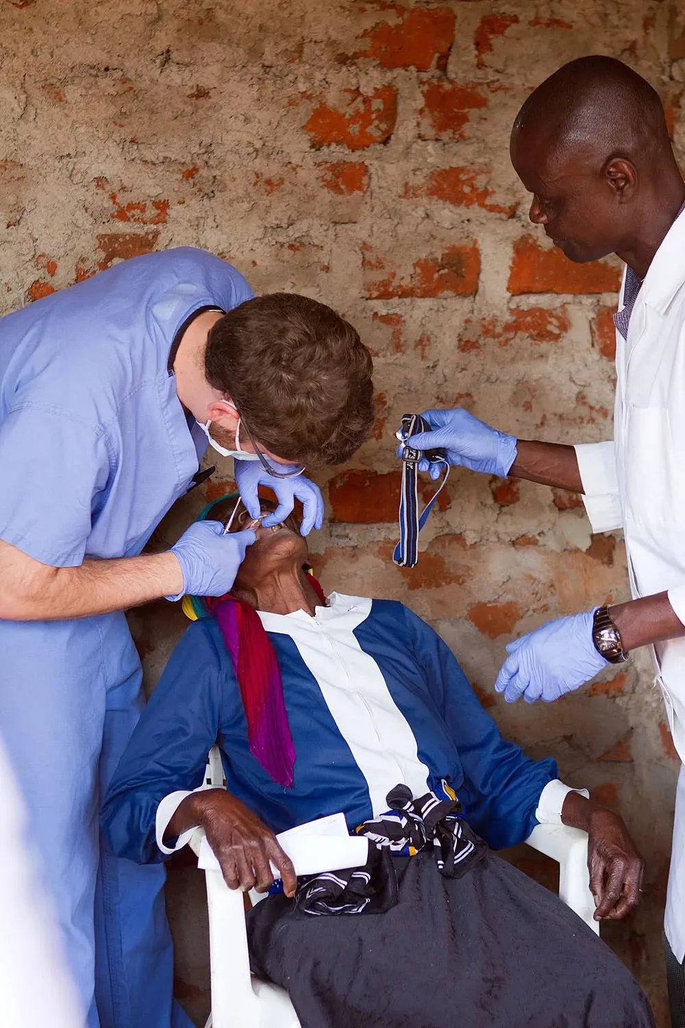 A healthcare worker in blue scrubs examines an elderly woman's mouth while another man in a white coat holds a wristwatch nearby, both wearing blue gloves, against a rough brick wall background.