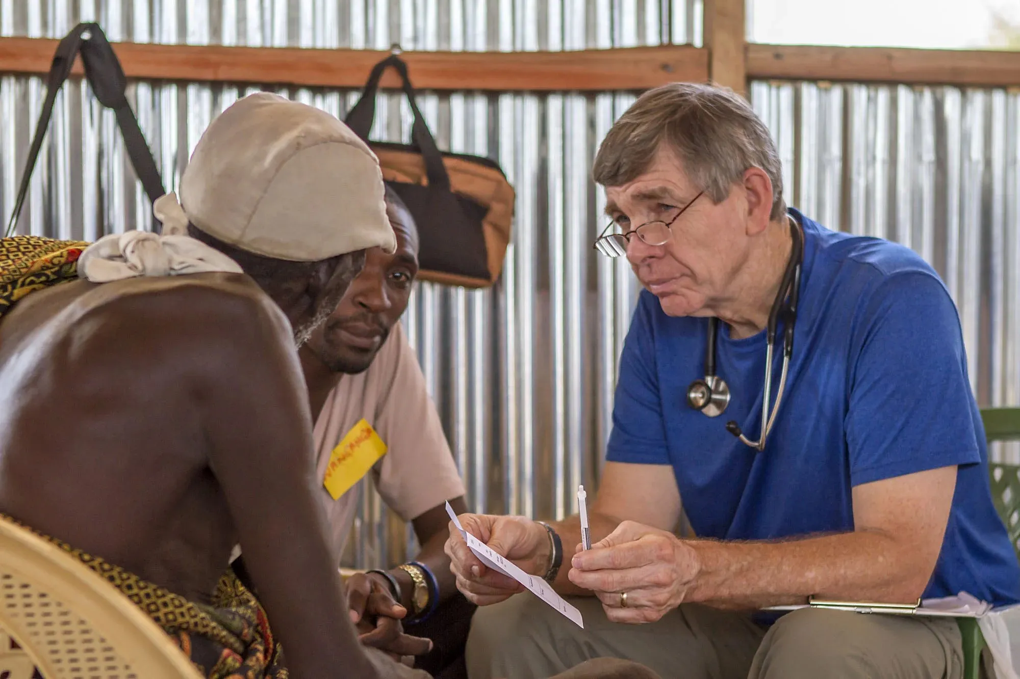 A doctor in a blue shirt with a stethoscope speaks with two men in a rustic room with corrugated metal walls.
