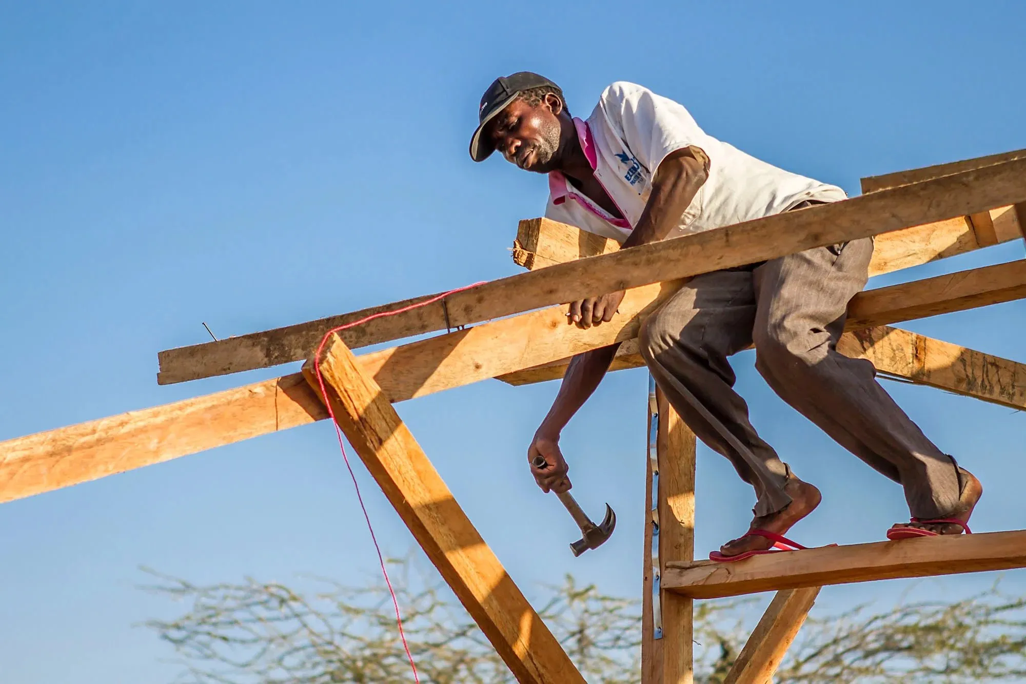 A man wearing a cap, white shirt, and brown pants working barefoot on a wooden roof frame using a hammer under a clear blue sky.