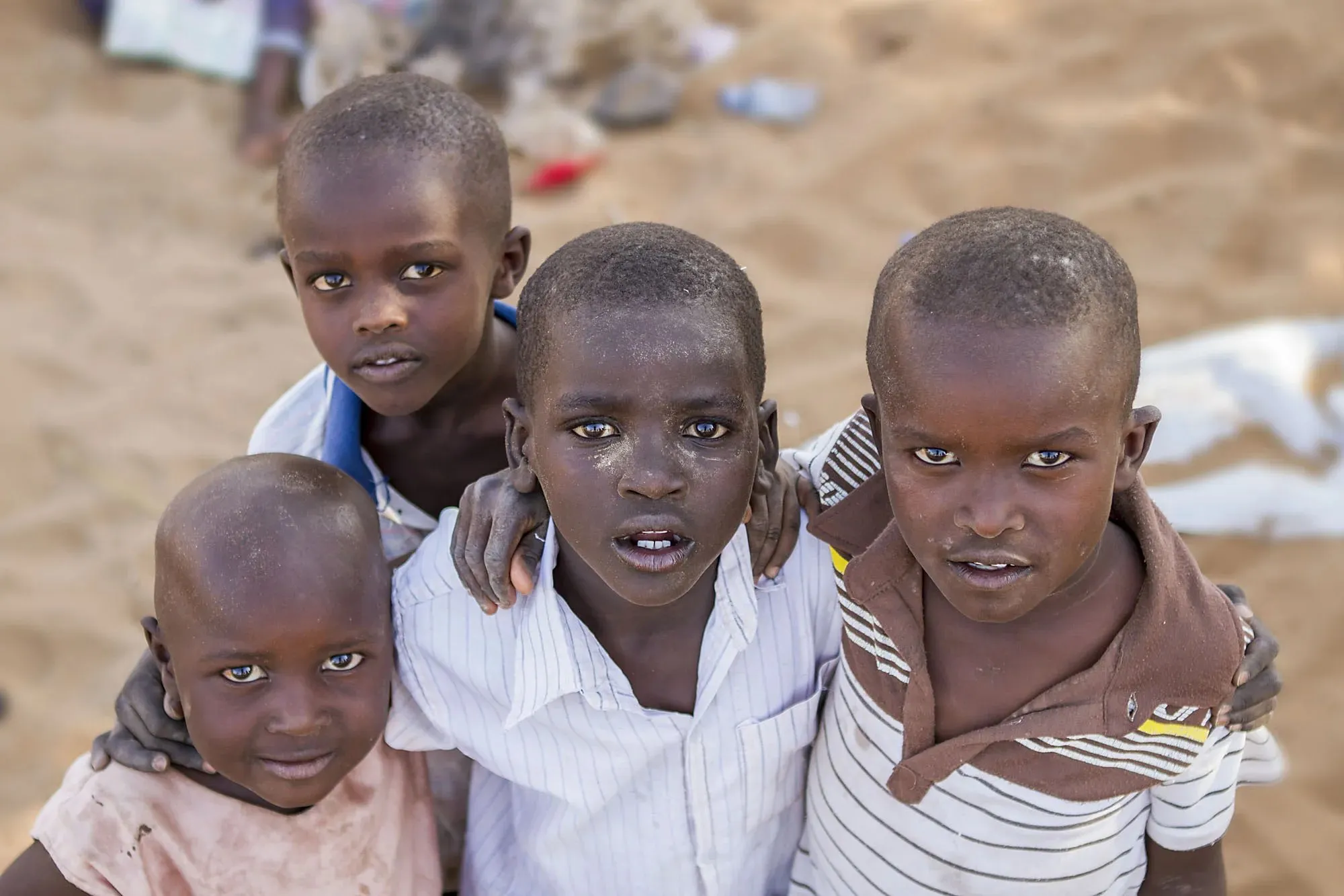 Four young boys with serious expressions standing close together outdoors on sandy ground.
