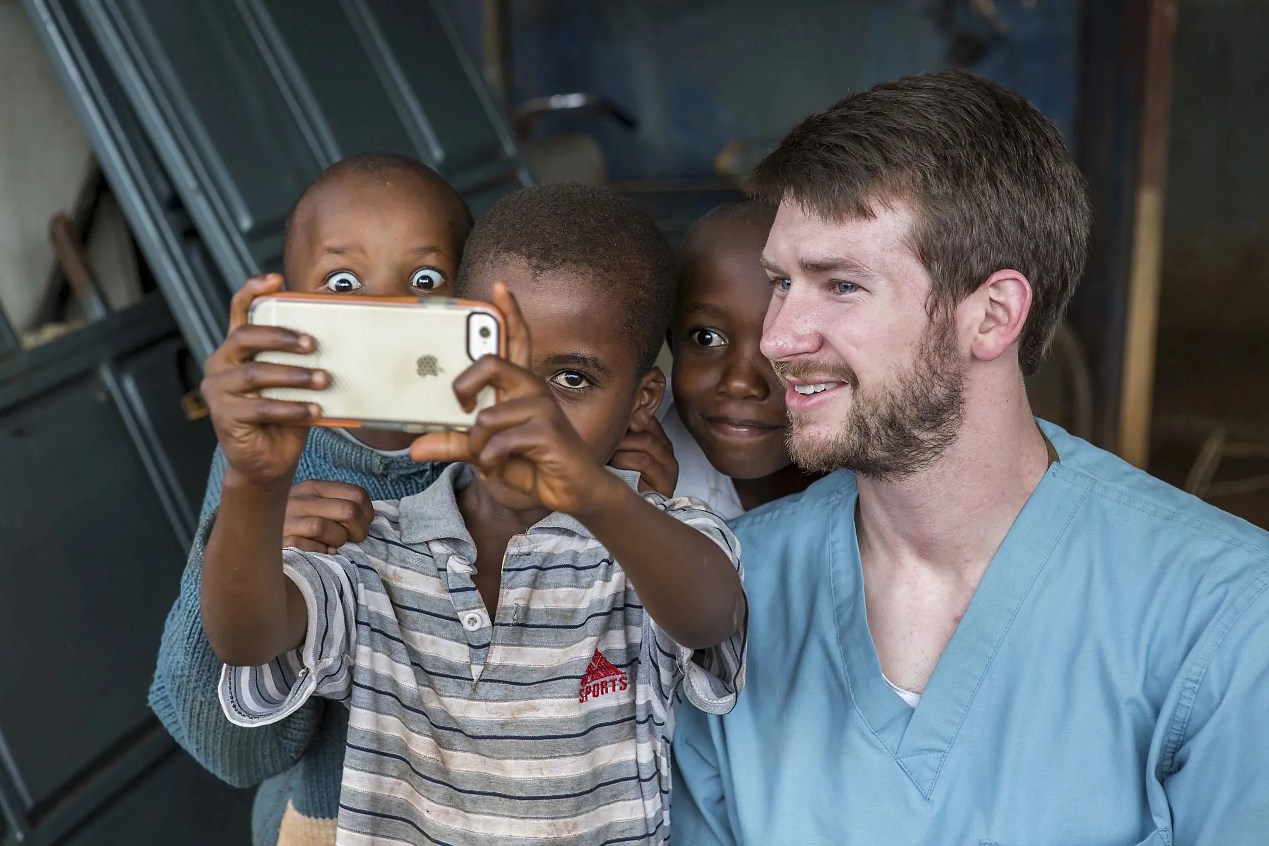 A smiling man in blue scrubs takes a selfie with three young boys, one of whom is holding the phone.