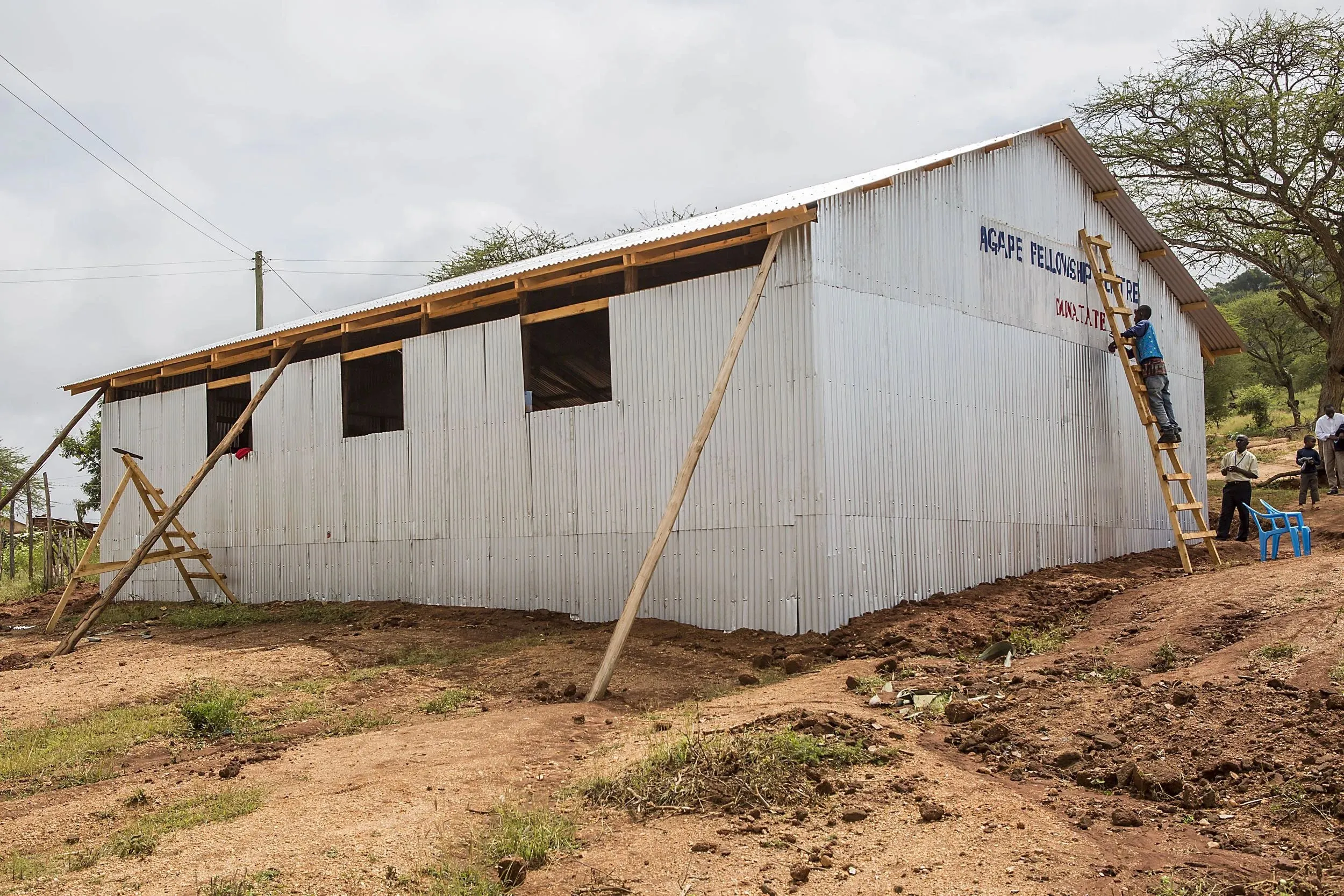 A man on a wooden ladder painting the words 'Agape Fellowship Centre' on the side of a large corrugated metal building supported by wooden beams.