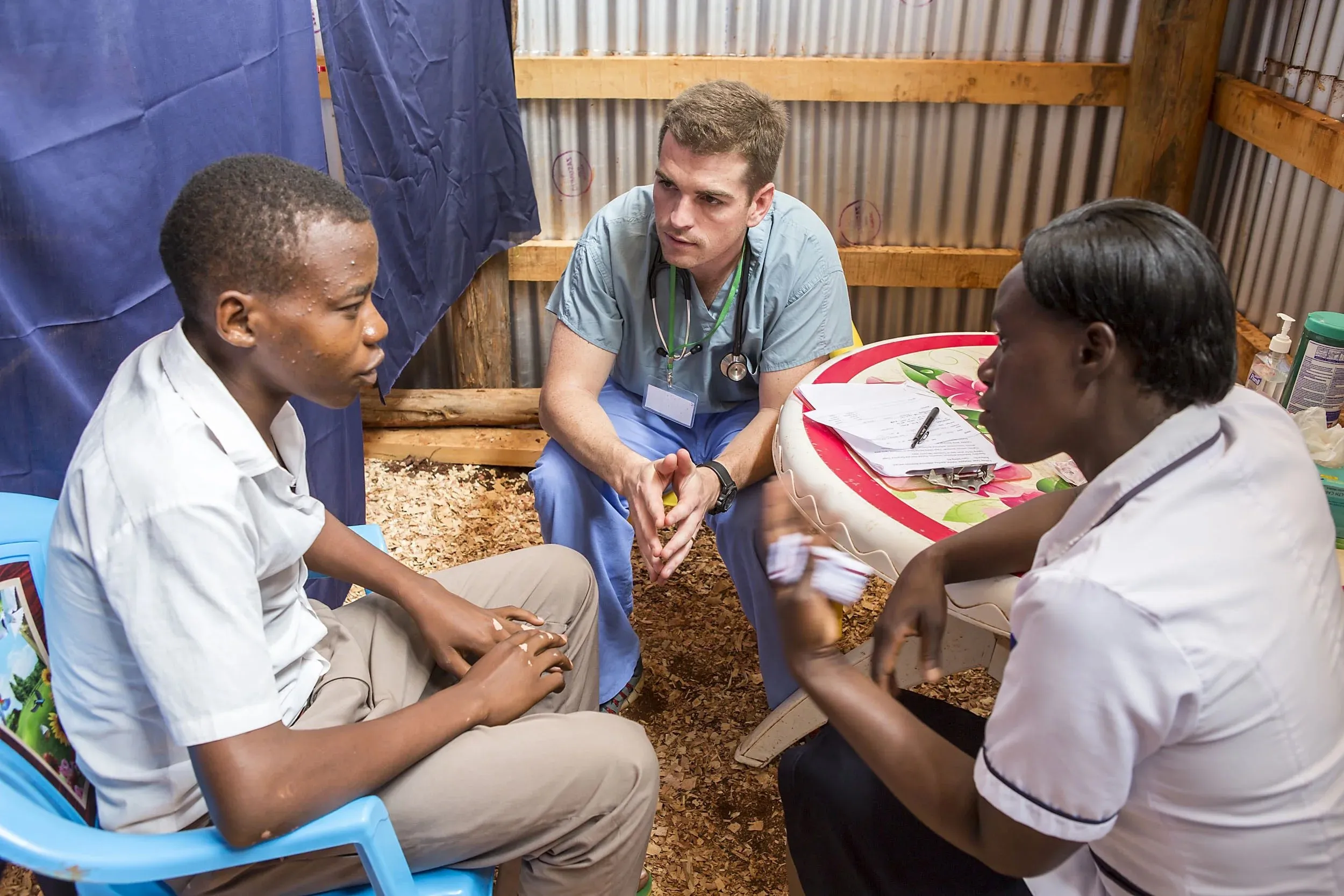 A male doctor in scrubs and stethoscope listening attentively to a young male patient and female nurse inside a rustic clinic room.