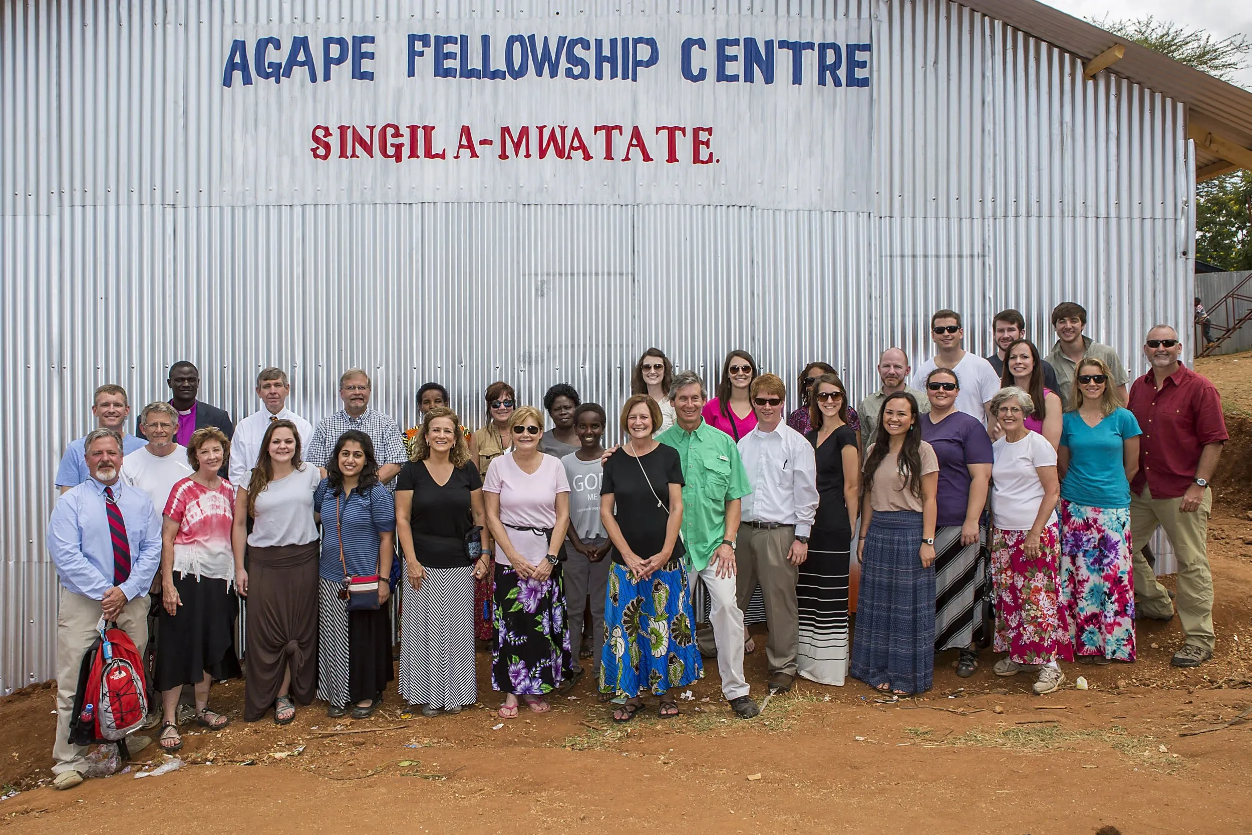 Group of diverse people standing and smiling in front of a corrugated metal building with the sign 'Agape Fellowship Centre Singila-Mwatate.'