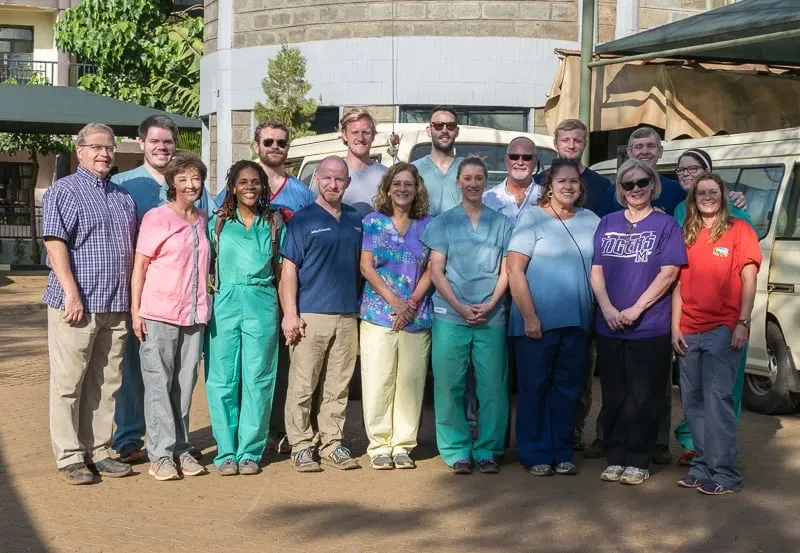 Group of fourteen healthcare professionals and volunteers standing outdoors in front of a medical vehicle and building.