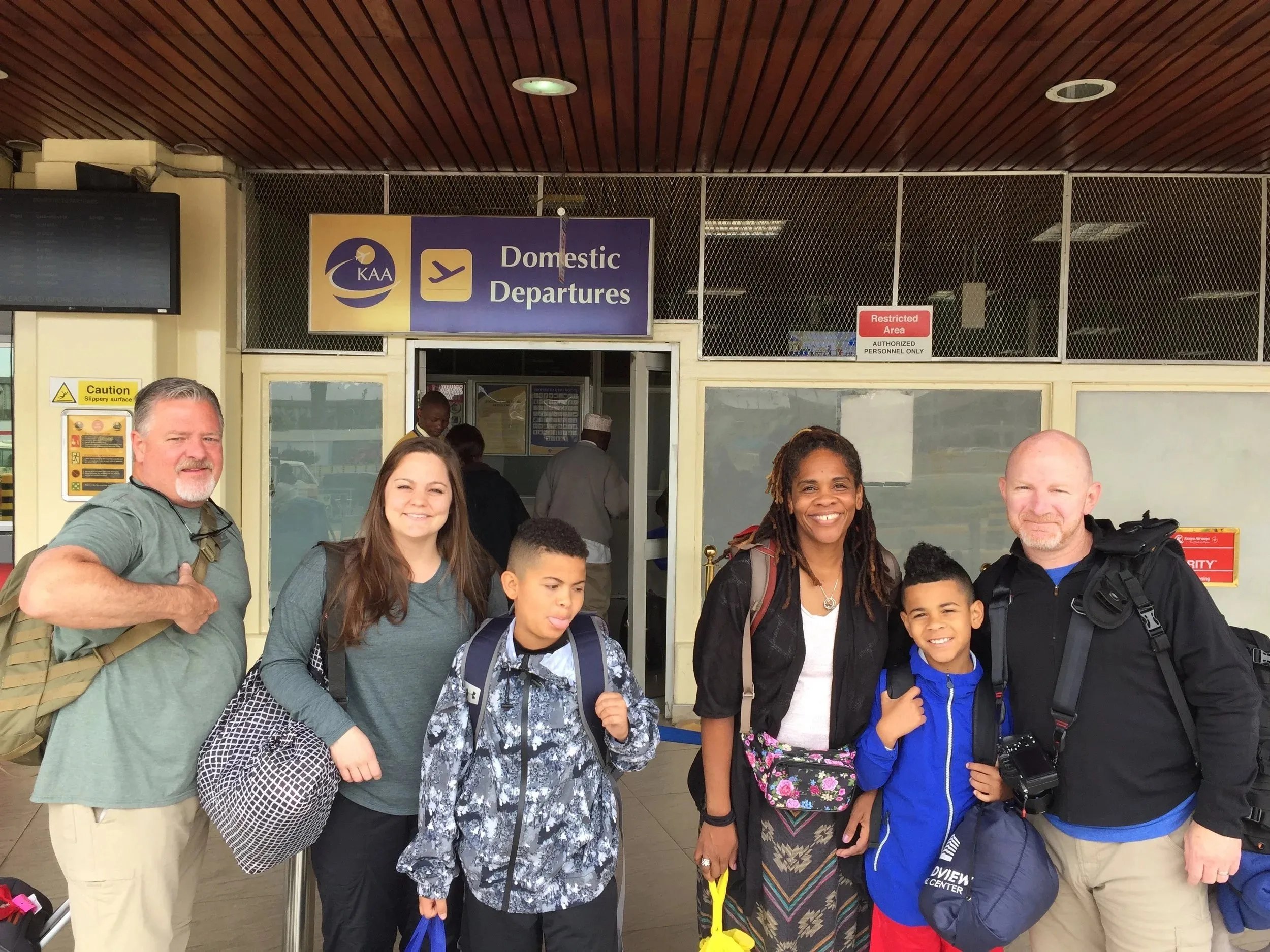 Group of six smiling travelers, including two children, standing outside a Domestic Departures entrance at an airport.