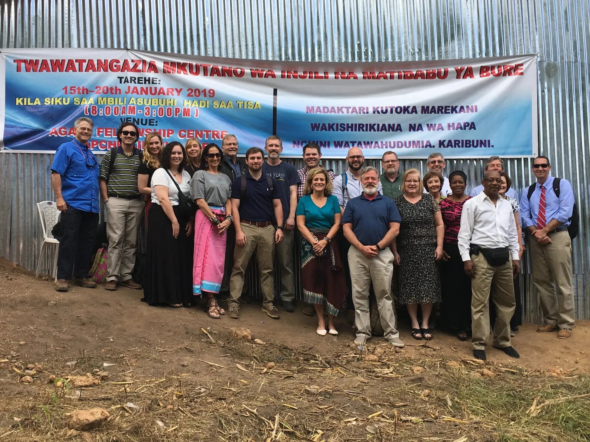 Group of diverse adults standing outdoors in front of a corrugated metal wall with a large banner advertising a January 2019 medical mission event.