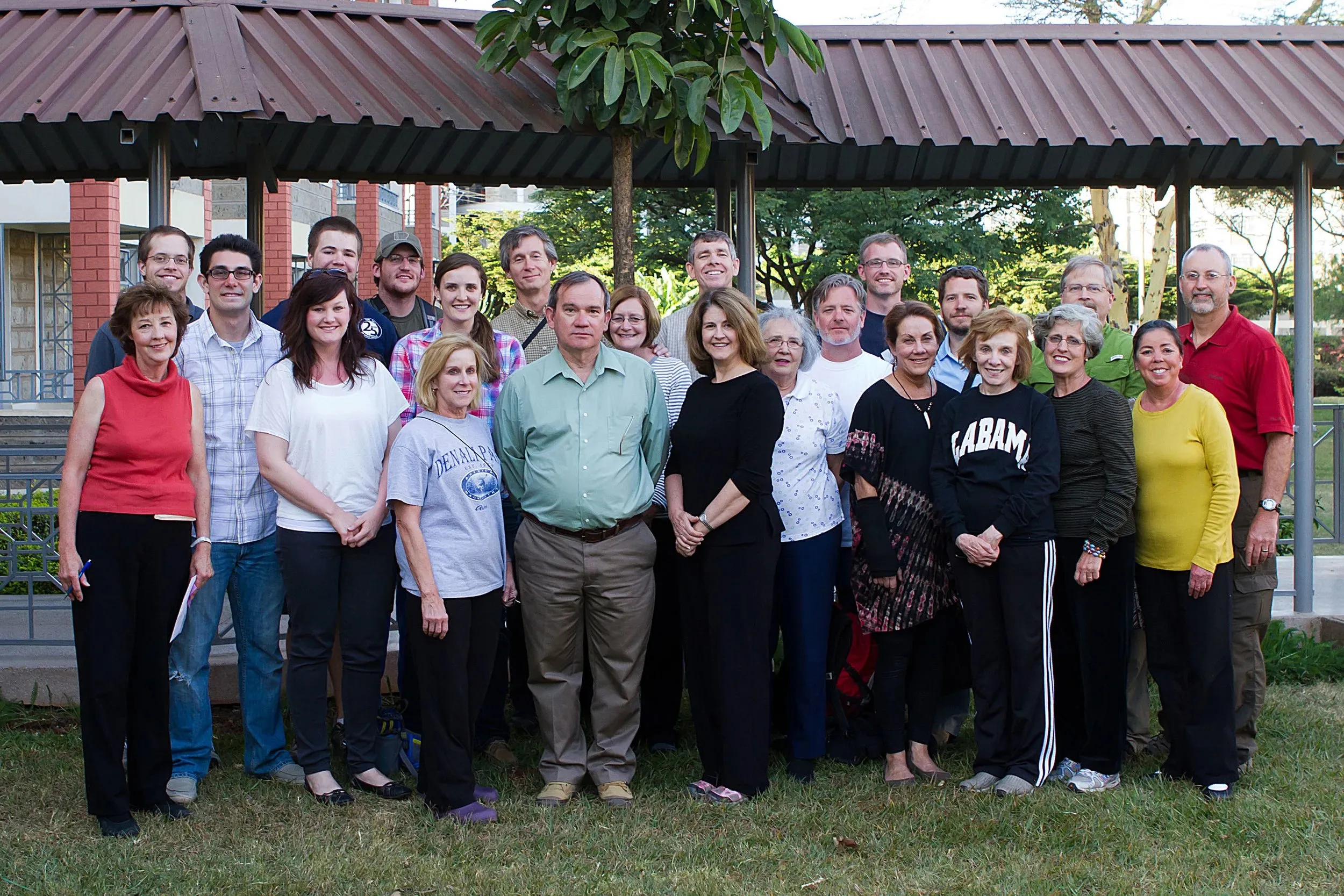 Group of diverse adults posing outdoors under a pavilion with trees and grass.