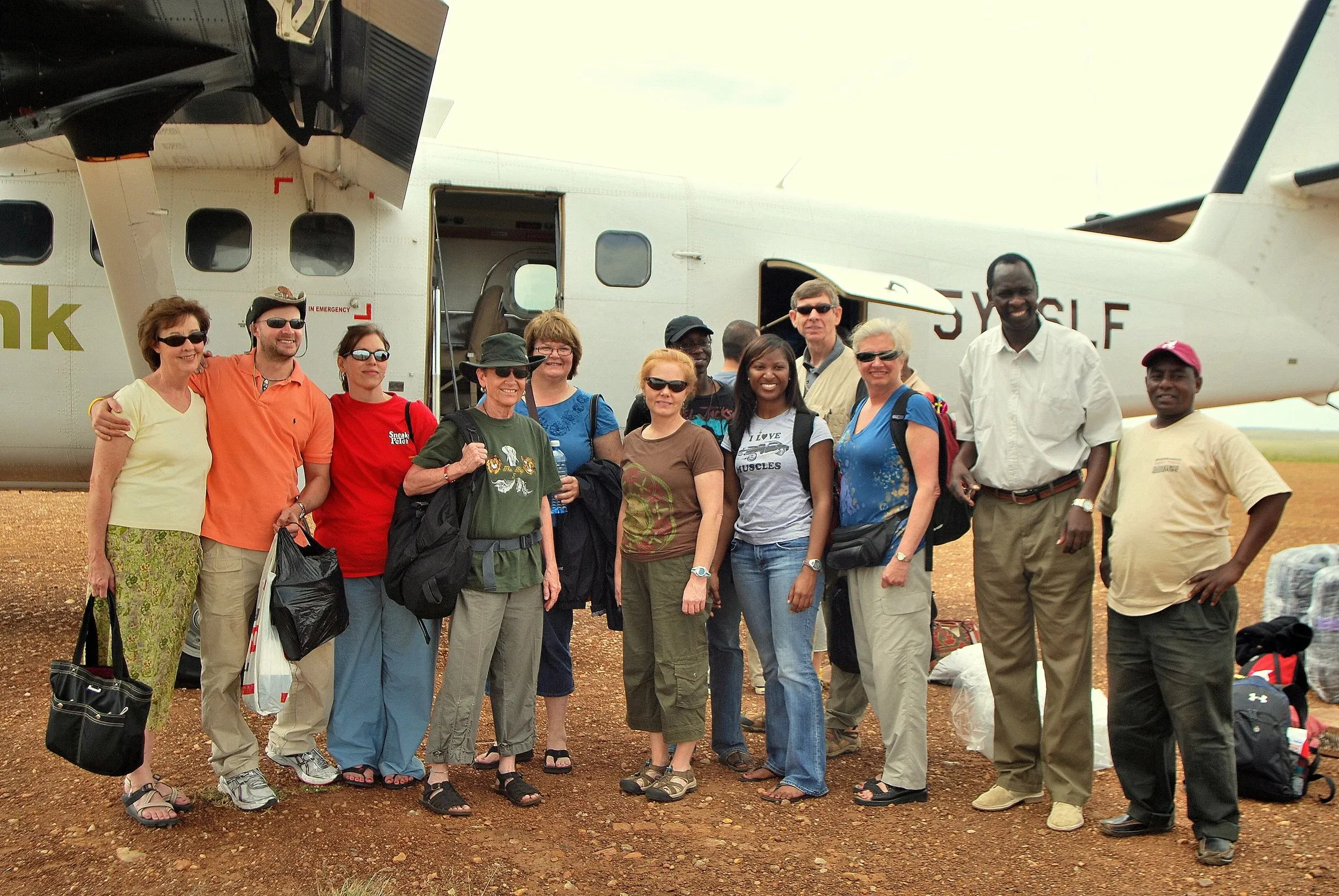 Group of diverse travelers posing together laughing near a small white airplane on a dirt airstrip.