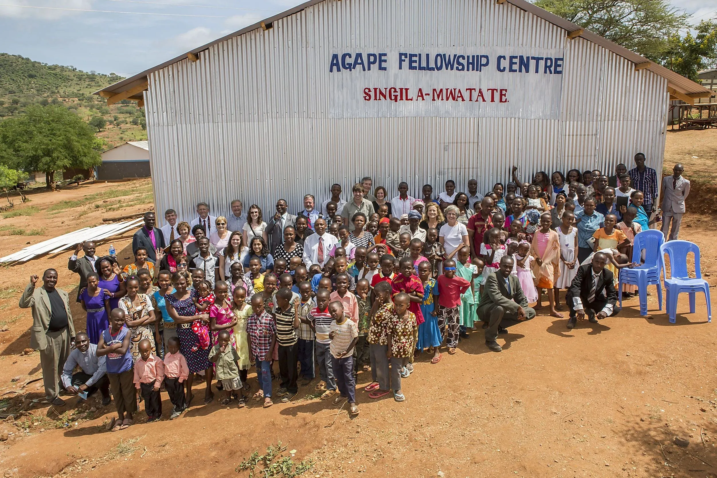 Large group of adults and children posing in front of a corrugated metal building with the sign 'Agape Fellowship Centre Singila-Mwatate' on a sunny day.