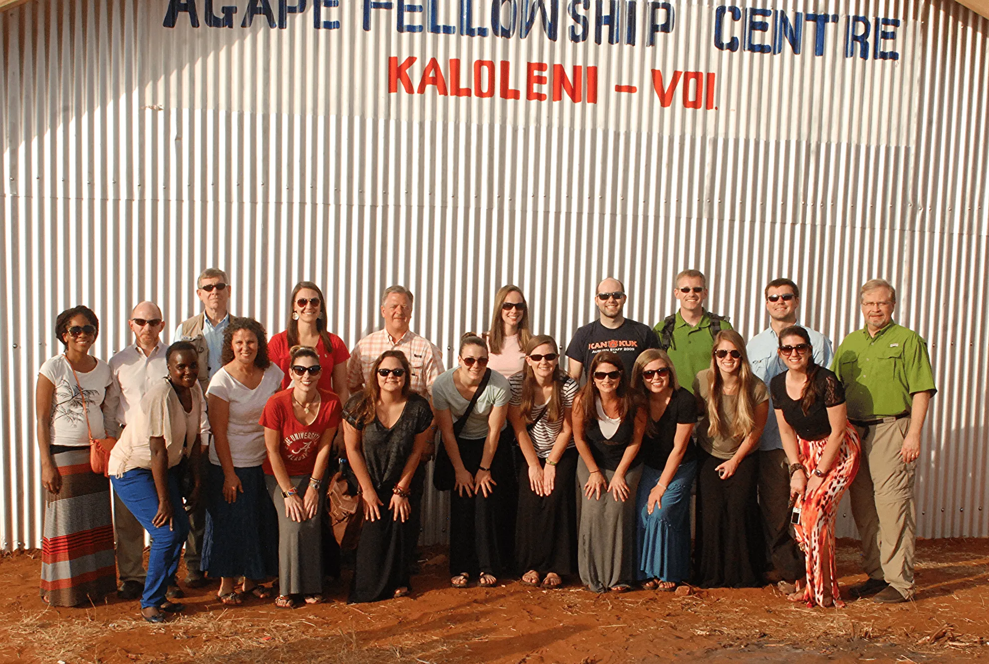 Group of smiling people standing and kneeling in front of a corrugated metal wall with 'AGAPE FELLOWSHIP CENTRE KALOLENI - VOI' sign.