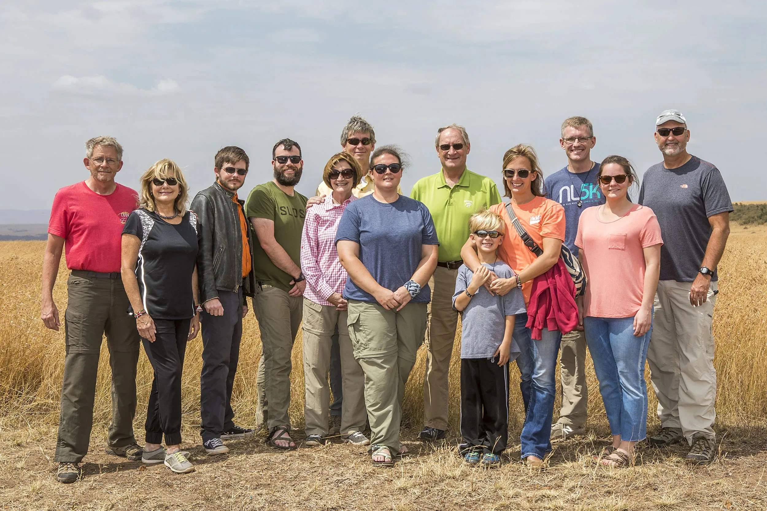 Group of thirteen people wearing casual outdoor clothing and sunglasses standing in a dry grassy field under a partly cloudy sky.