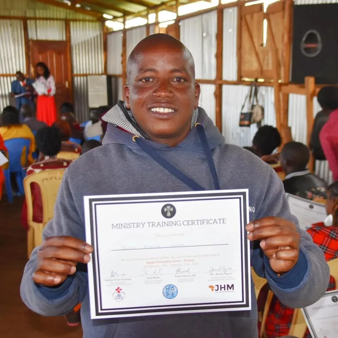 Man smiling and holding a Ministry Training Certificate inside a room with other seated people.