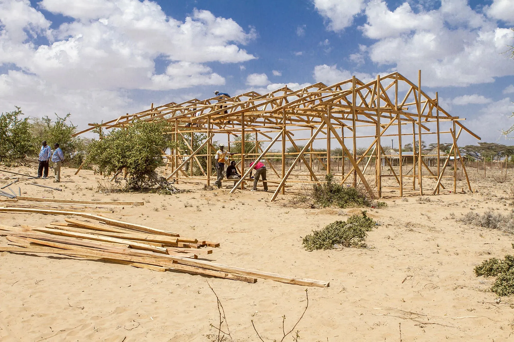 Several workers assembling a large wooden framework structure on a sandy, arid landscape under a partly cloudy sky.