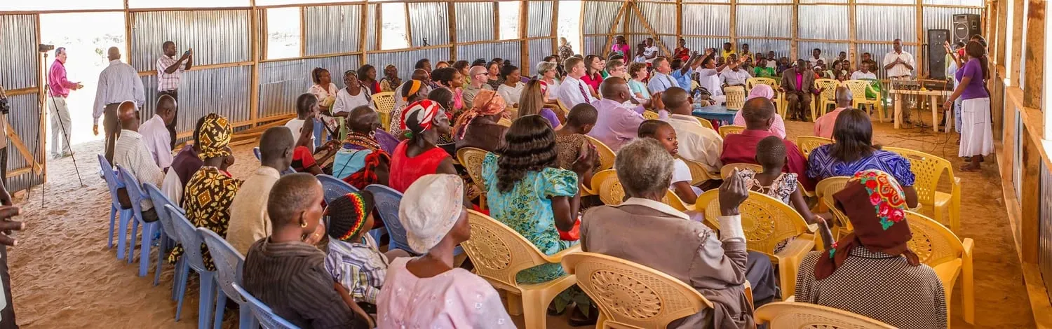 Community gathering inside a large open-sided wooden structure with people seated on plastic chairs listening to a speaker at the front.