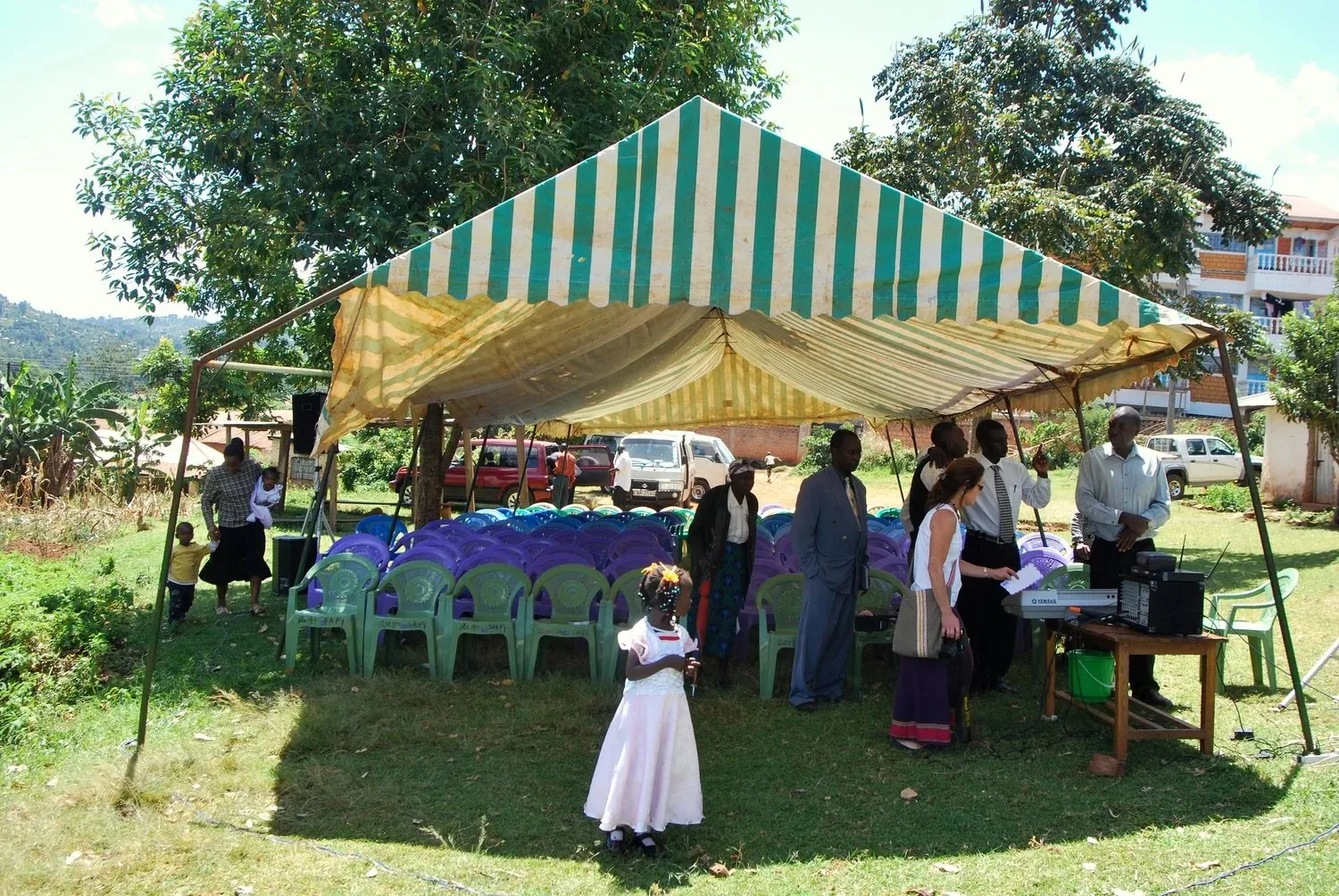 Outdoor event with green and white striped tent, rows of green and purple chairs, and a group of people conversing beside a keyboard and speaker setup.