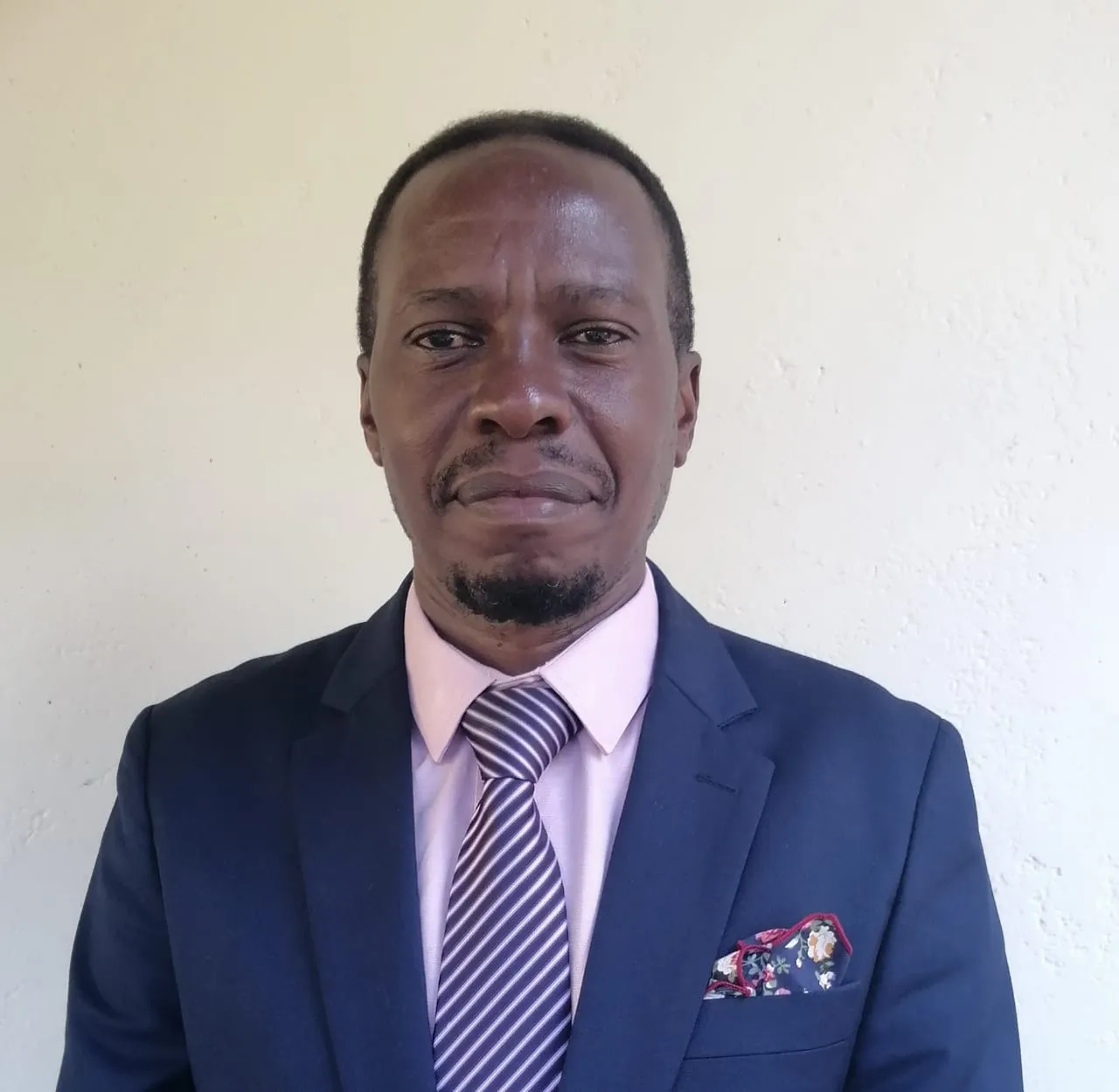 Man wearing a navy blue suit jacket, pink shirt, striped purple tie, and floral pocket square, standing against a plain light wall.