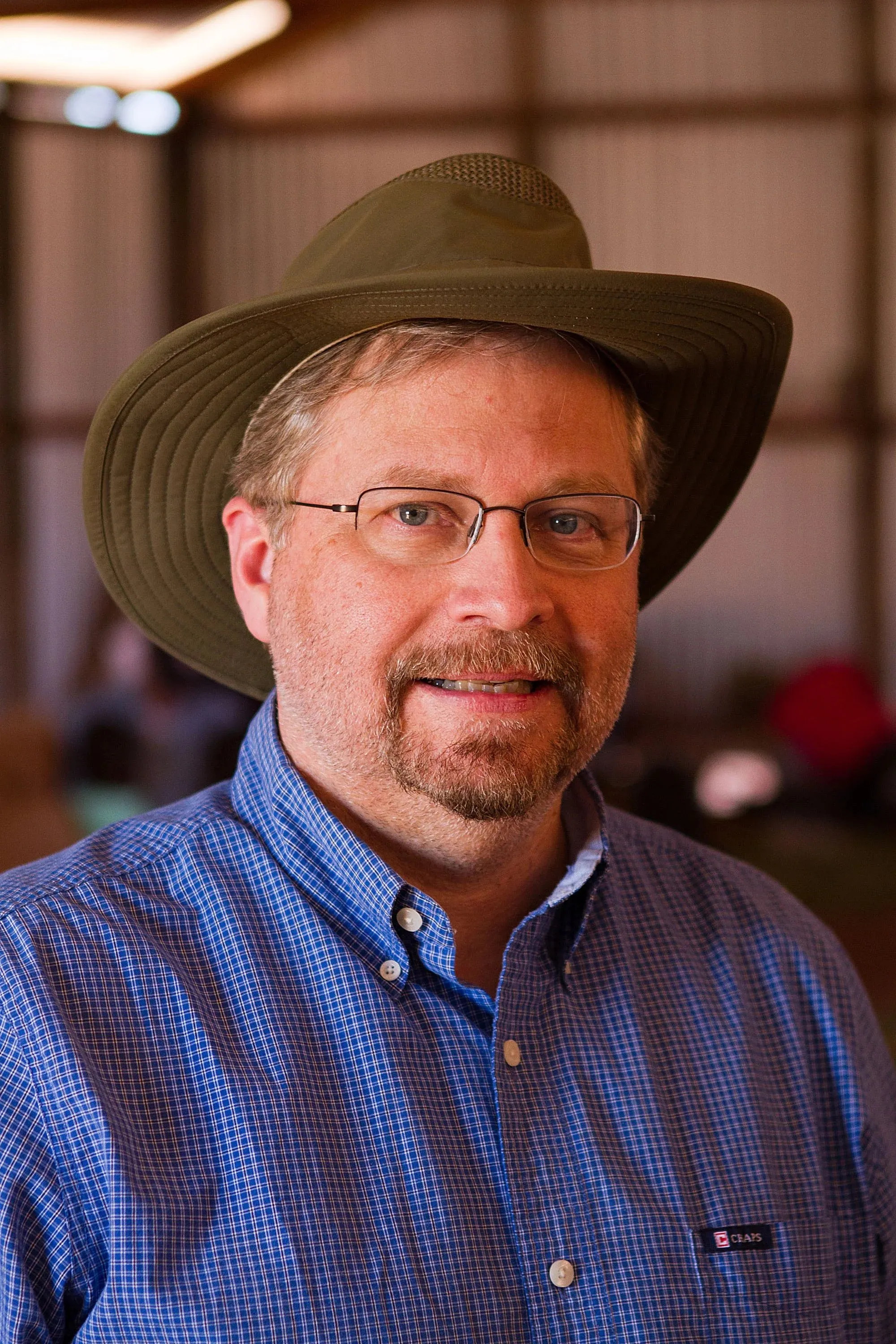 Middle-aged man wearing glasses, a green wide-brimmed hat, and a blue checkered button-up shirt smiling indoors.