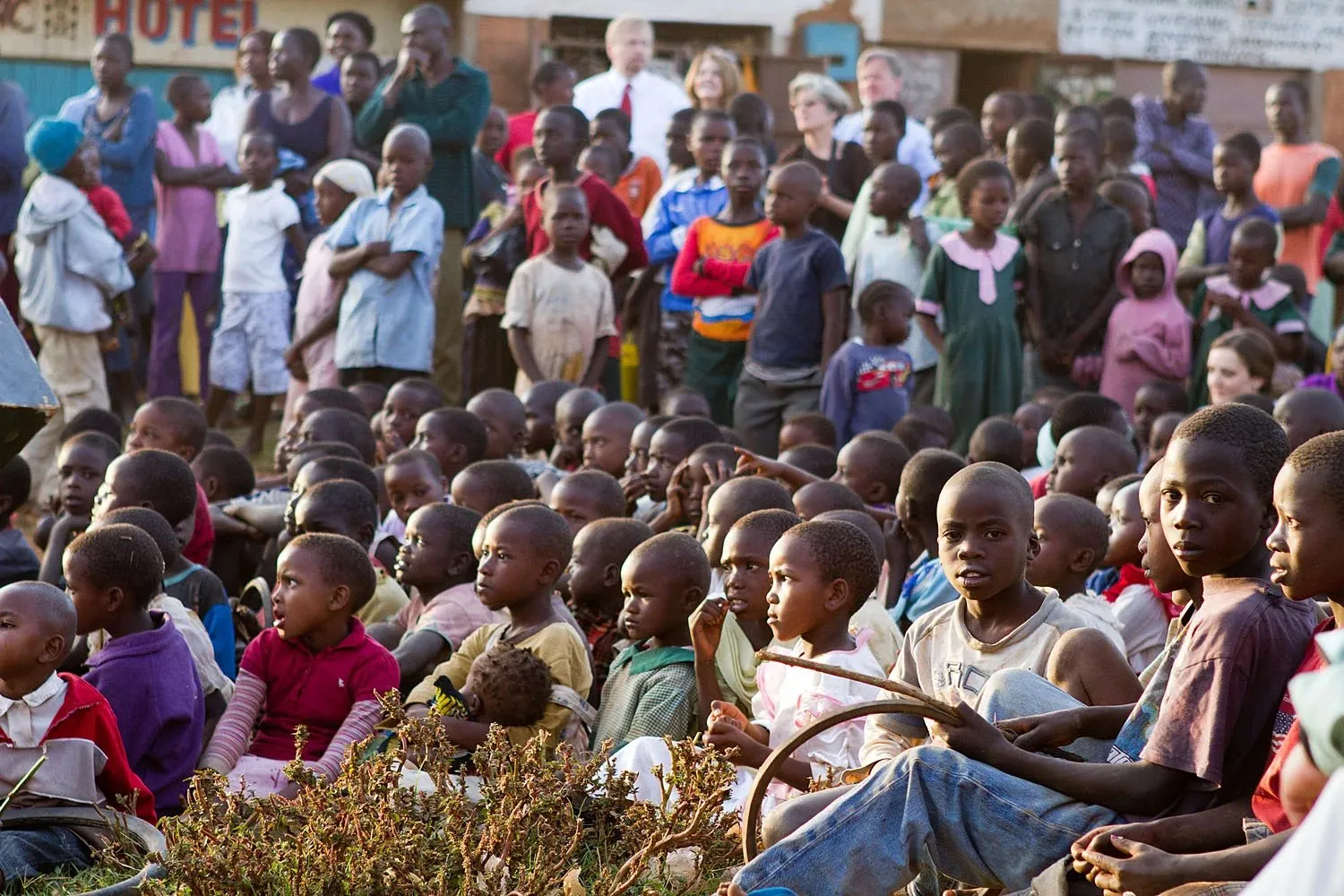 Large group of children and a few adults gathered outdoors with some children seated on the ground and others standing in the background.