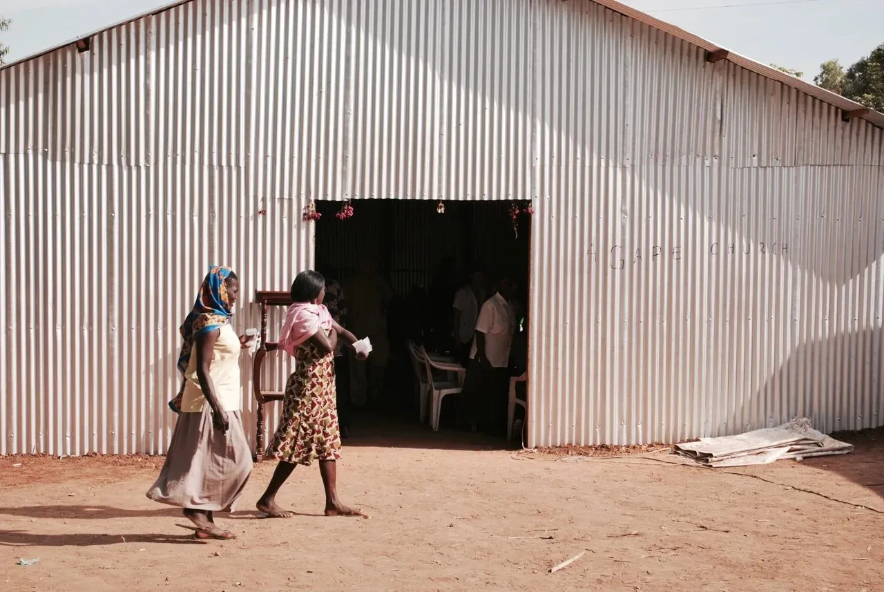 Two women walking barefoot on dirt ground outside a corrugated metal building with an open entrance that reveals people seated inside.
