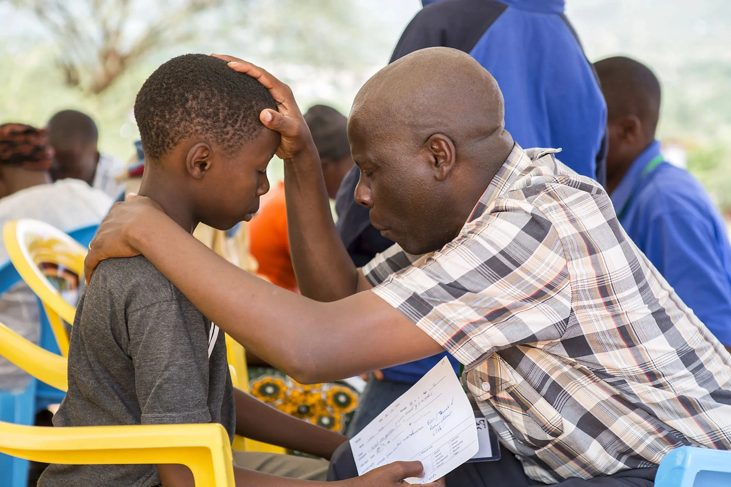Man in a plaid shirt comforting a young boy by placing his hand on the boy’s head while the boy sits with eyes closed holding a paper.