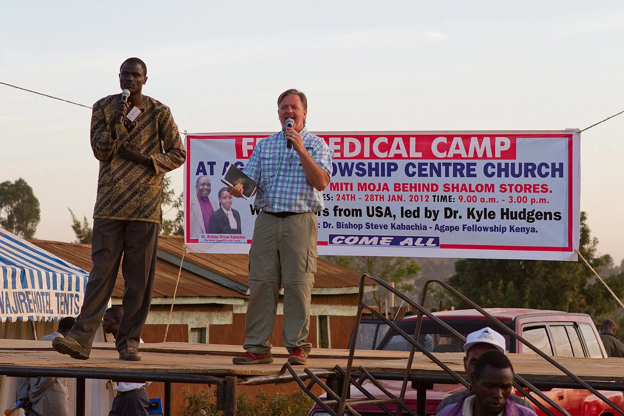 Two men speaking into microphones on an outdoor stage in front of a banner advertising a free medical camp at Agape Fellowship Centre Church.