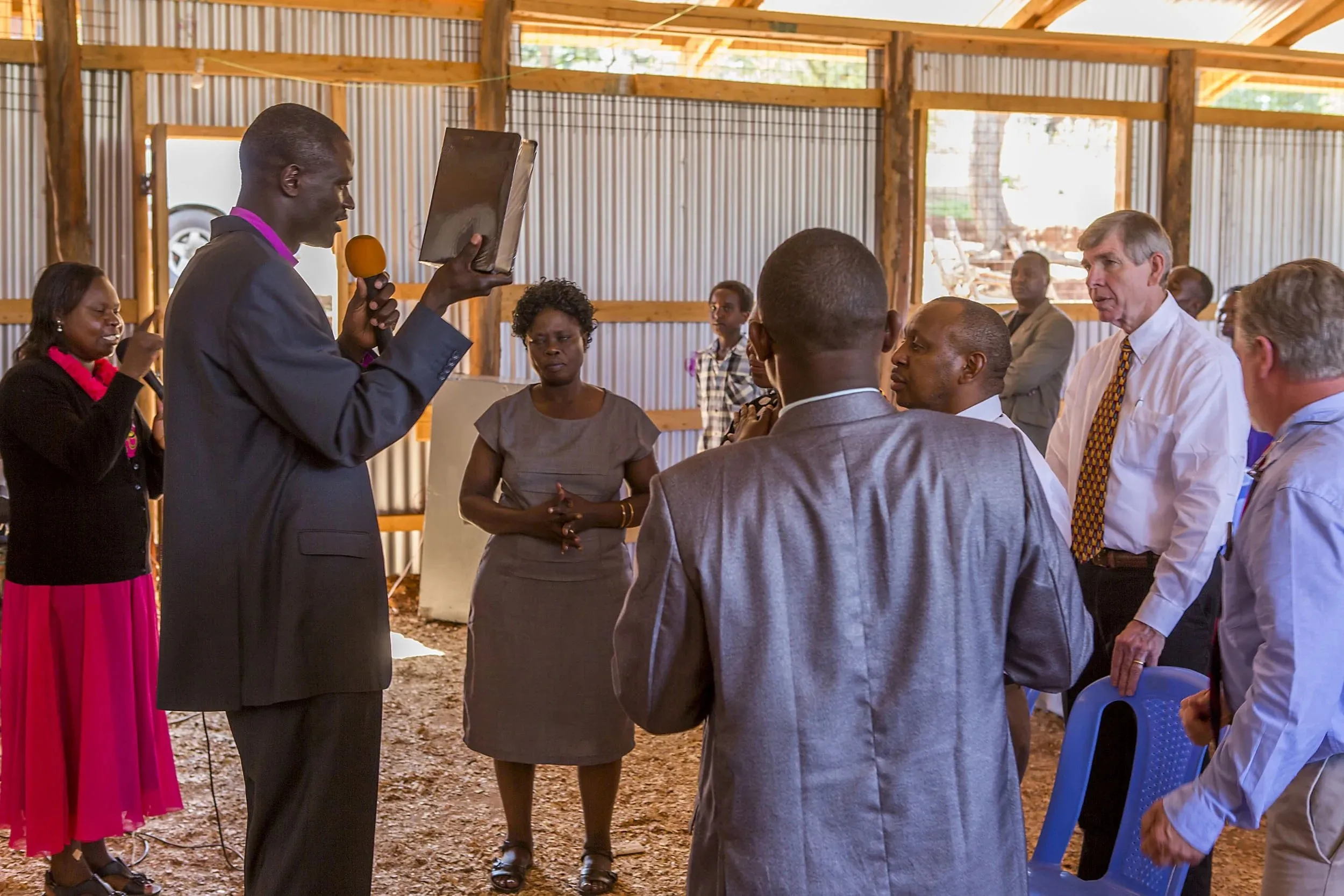 Man in a dark suit holding a microphone and a book, speaking to a group of people standing inside a wooden and metal room.