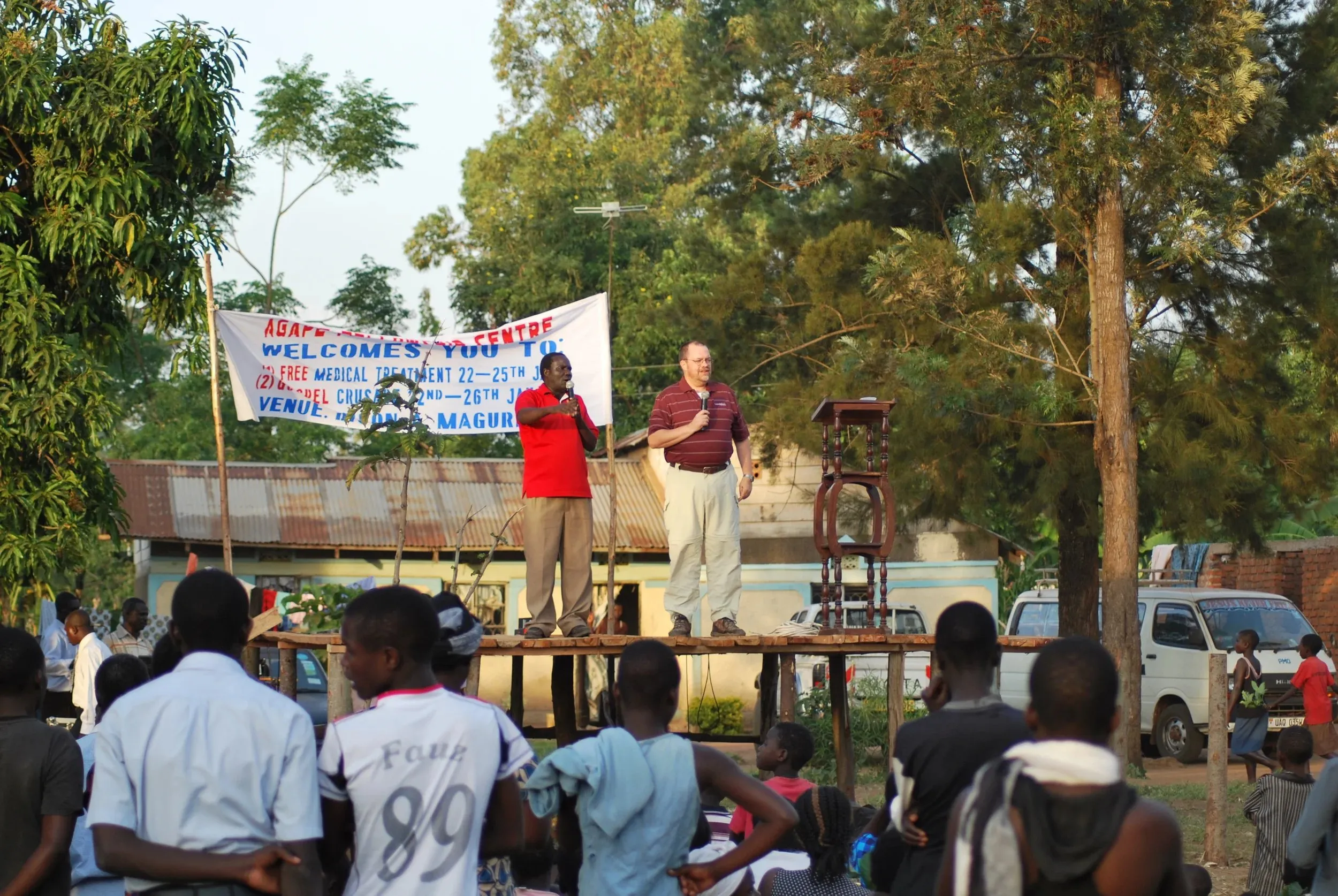 Two men on a wooden platform speaking to a crowd outdoors with a banner behind them and trees around.