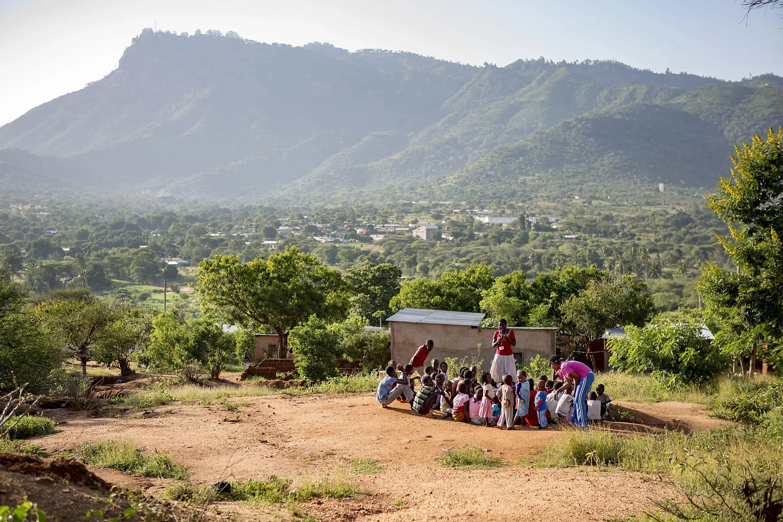 Group of children sitting and standing on dry ground near two adults outdoors with a mountainous landscape in the background.