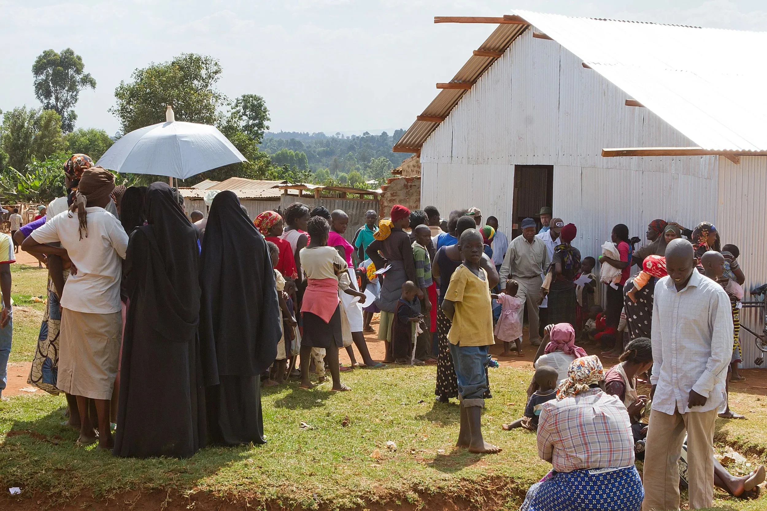 Large group of people, including women, children, and men, gathered outside a white building in a rural setting.
