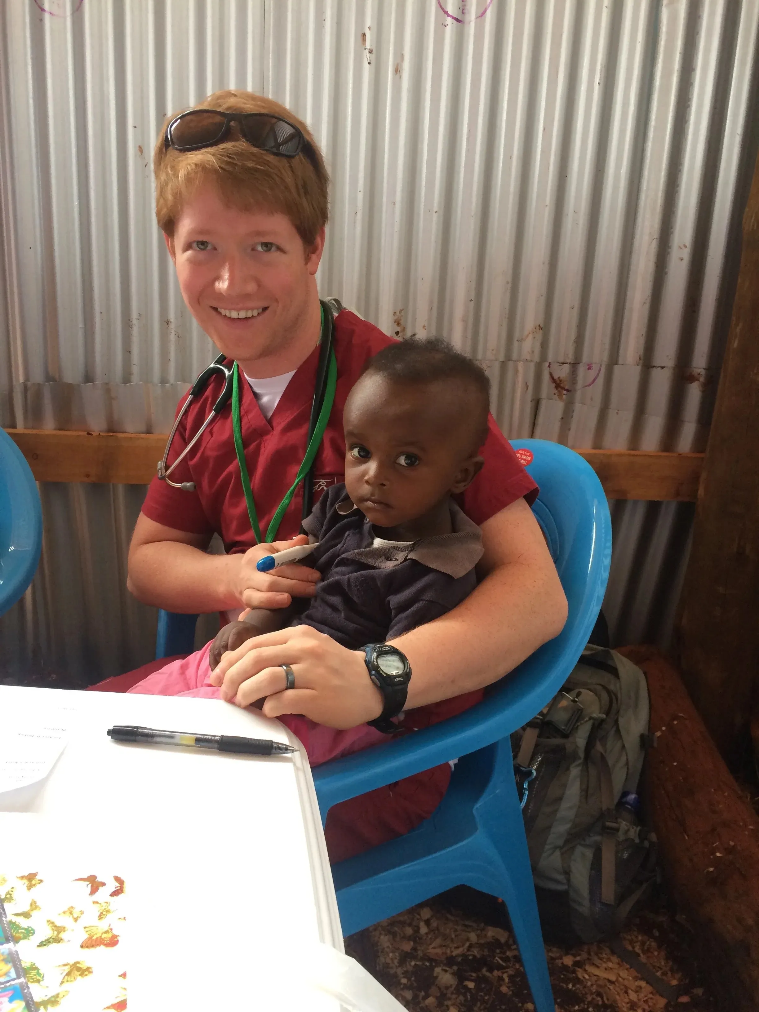 A smiling male healthcare worker in red scrubs holding a young child sitting on his lap, inside a room with corrugated metal walls.