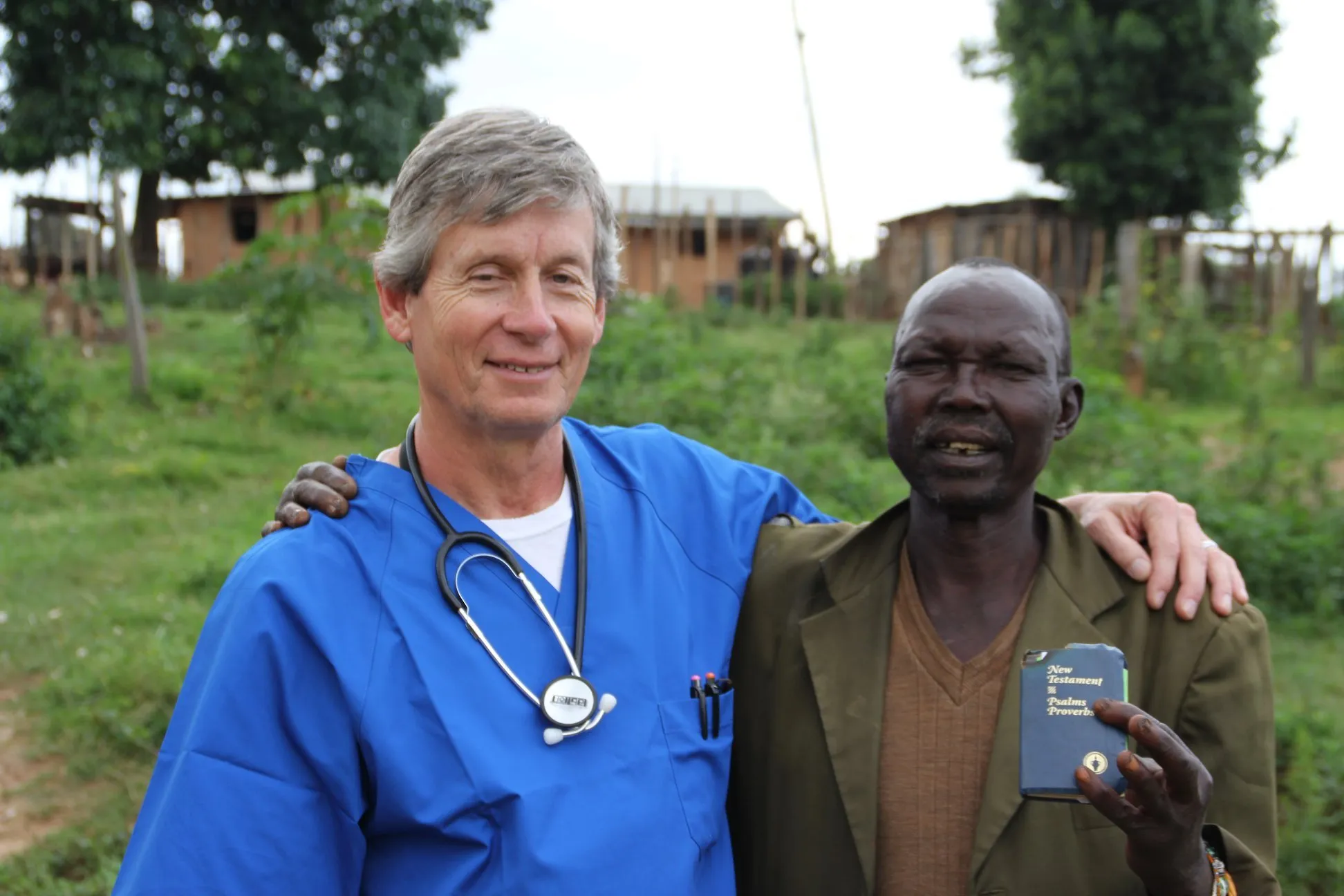 A smiling male doctor in blue scrubs with a stethoscope embraces an older man holding a small Bible outdoors.