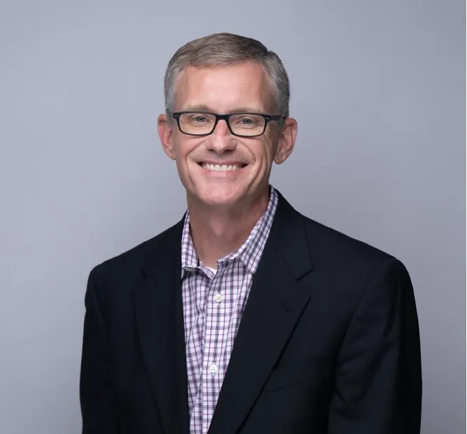 Middle-aged man wearing glasses, a checkered shirt, and a black blazer smiling against a plain gray background.