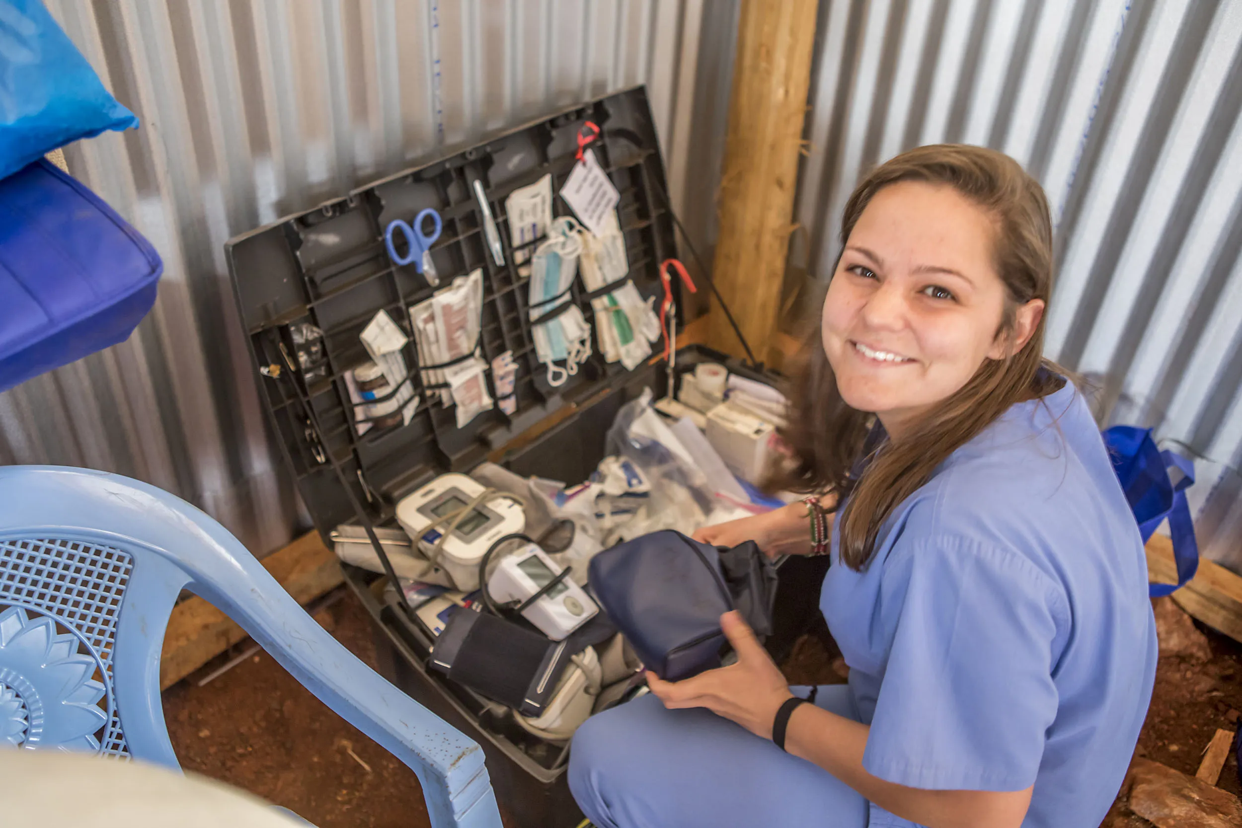 Smiling woman in blue scrubs organizing medical supplies from an open kit inside a corrugated metal structure.