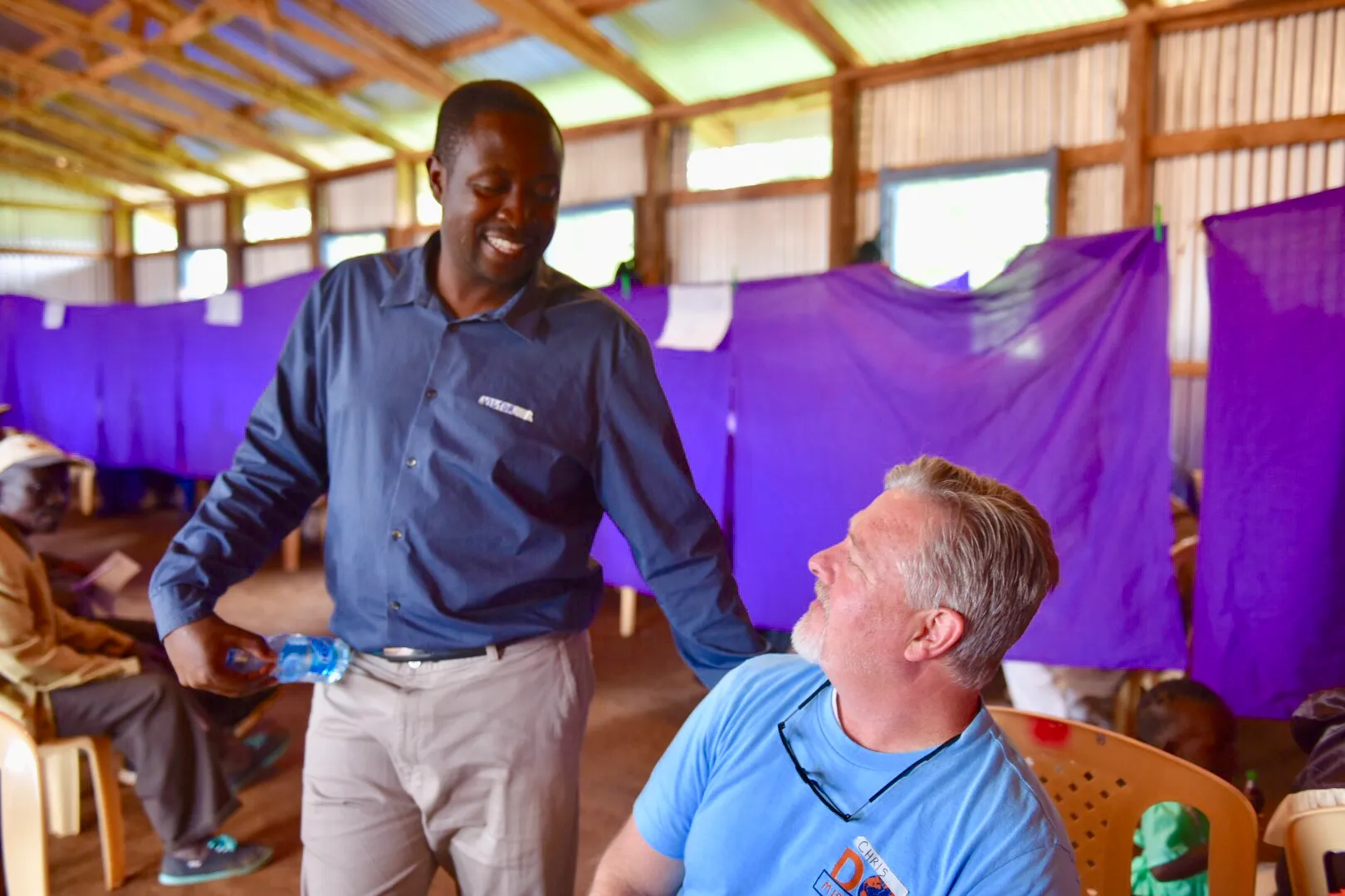 A smiling man in a blue shirt handing a water bottle to a seated man with gray hair wearing a blue T-shirt inside a building with purple curtains.