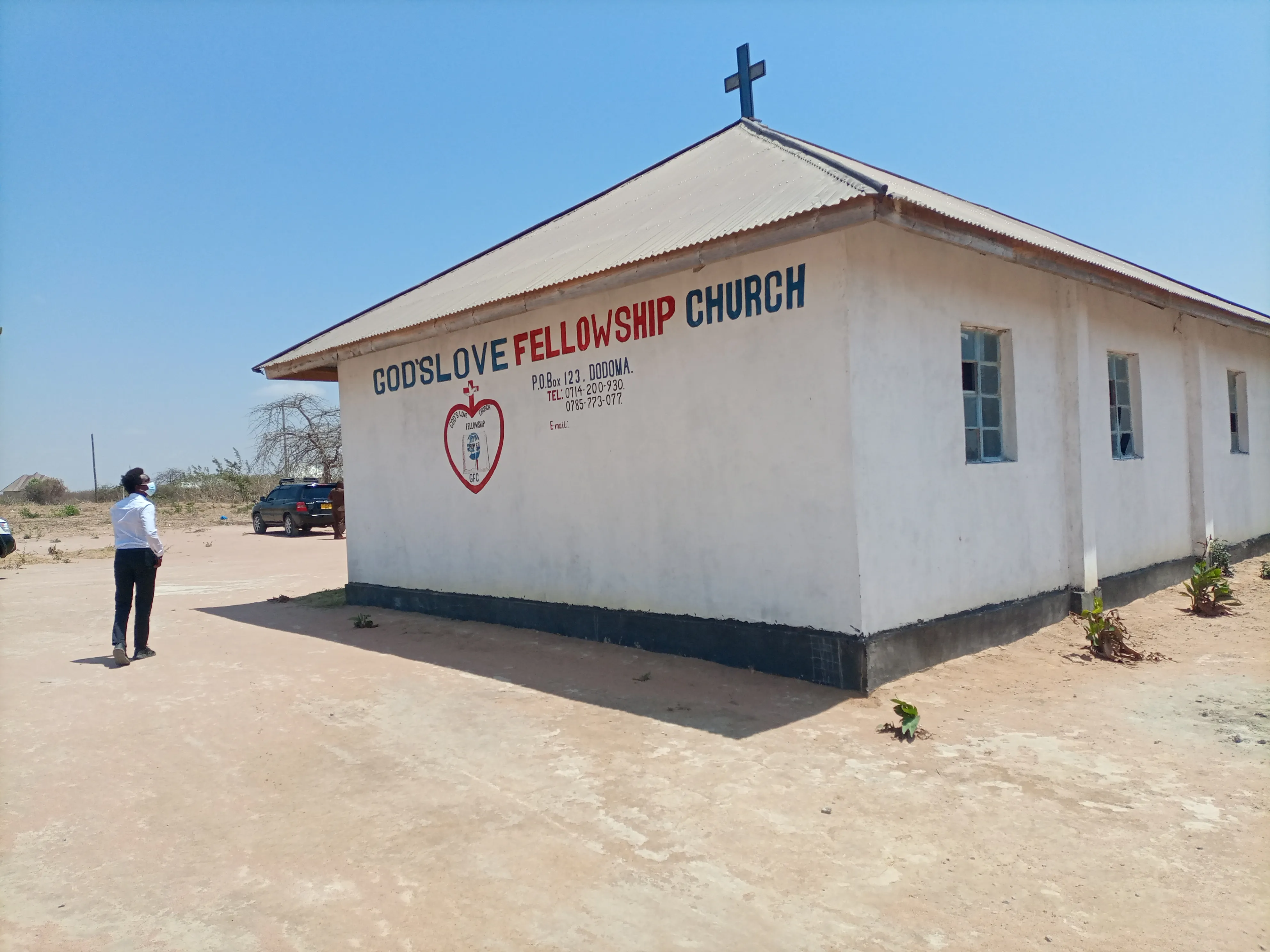 White church building with a cross on the roof and the sign 'God's Love Fellowship Church' painted on the side, with a man wearing a mask walking nearby.