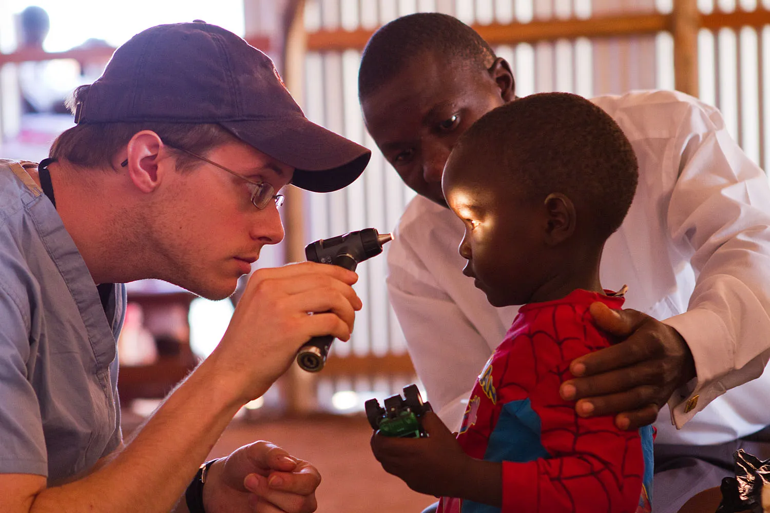 Doctor using an otoscope to examine a young boy's eye while the boy's guardian supports him.